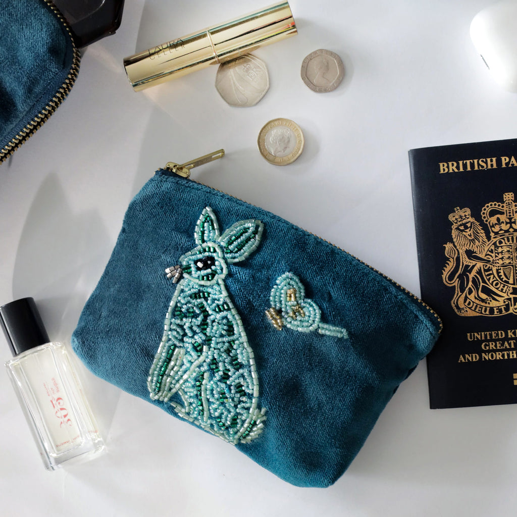 Teal embroidered pouch with hare design on a white surface with cosmetic items and a passport.