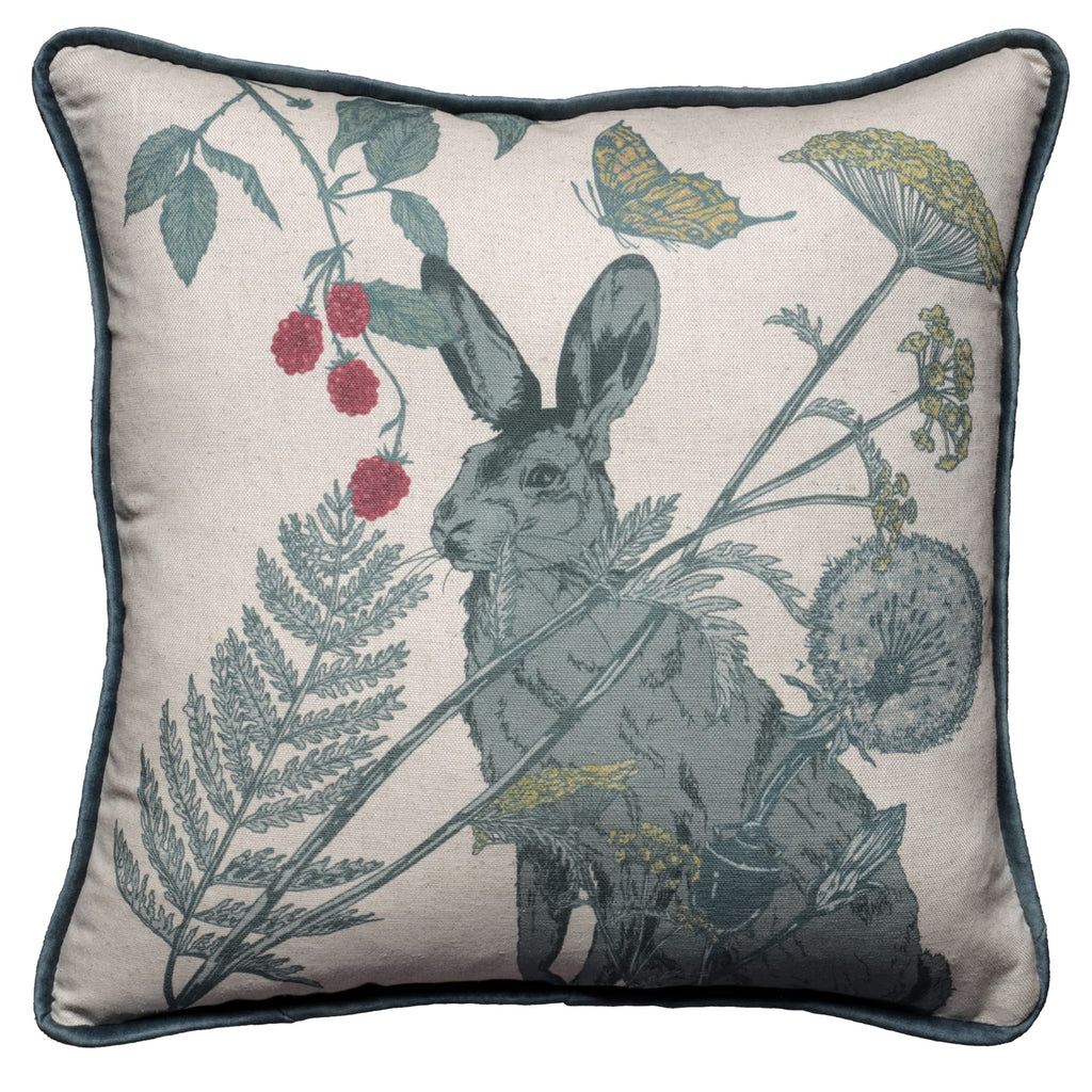 Square cushion featuring a detailed illustration of a hare surrounded by wildflowers, ferns, dandelions, and red berries. The design includes a butterfly in flight on a natural linen-coloured background, with soft blue velvet piping around the edges for a refined finish.