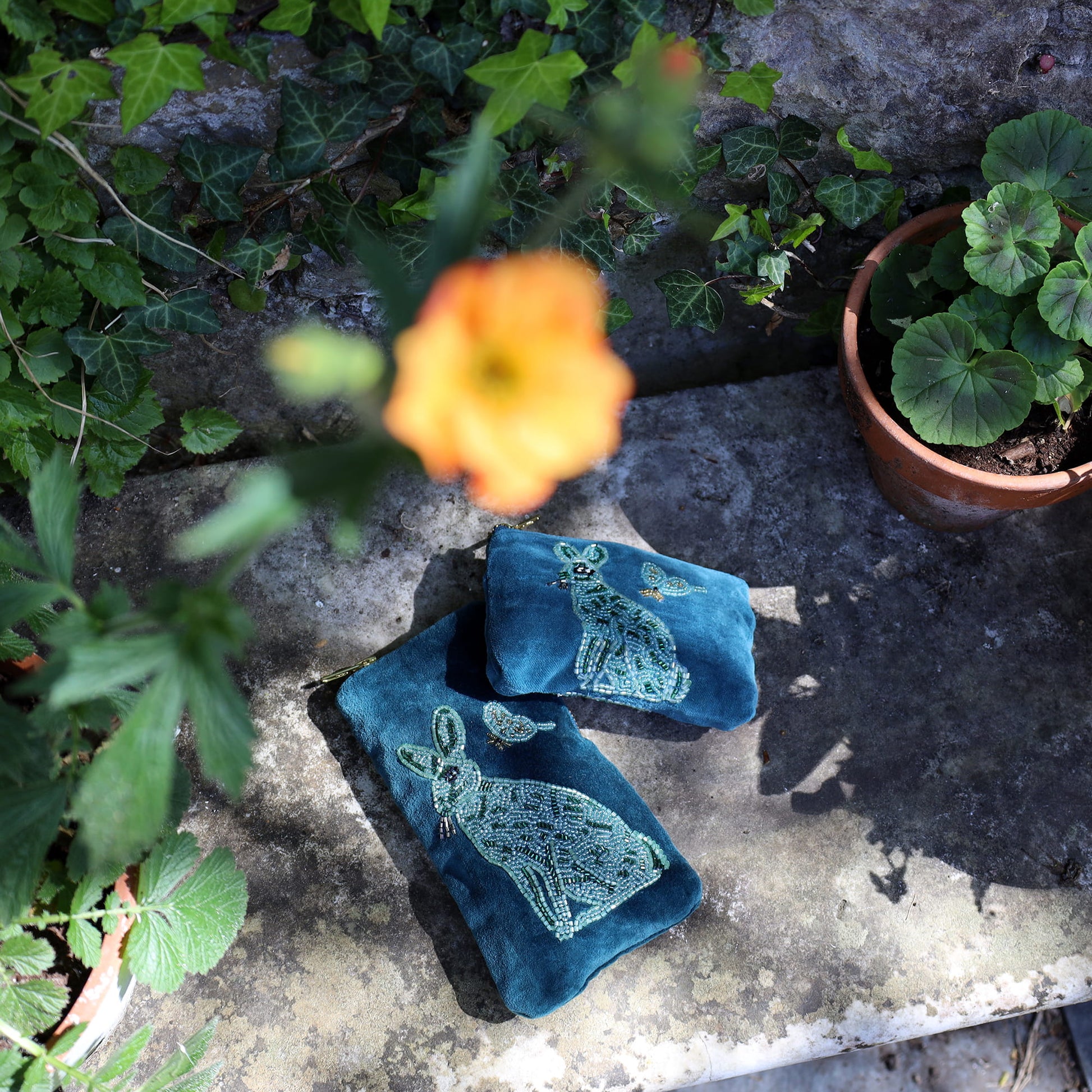 Blue embroidered fabric with cat designs on a stone surface surrounded by plants and flowers.