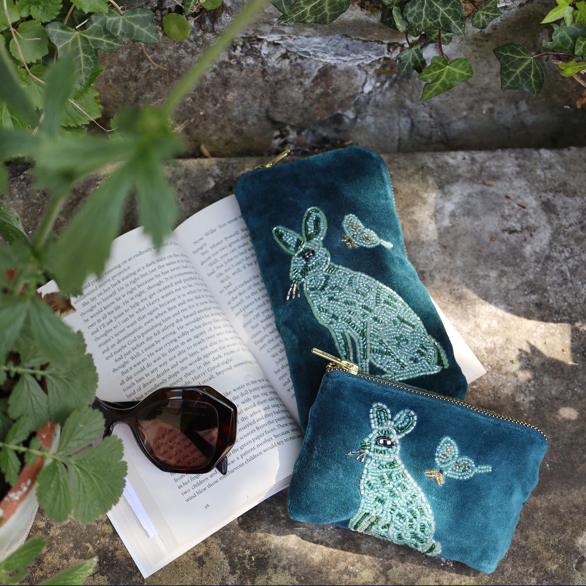 Two blue embroidered pouches on an open book with sunglasses, surrounded by greenery.