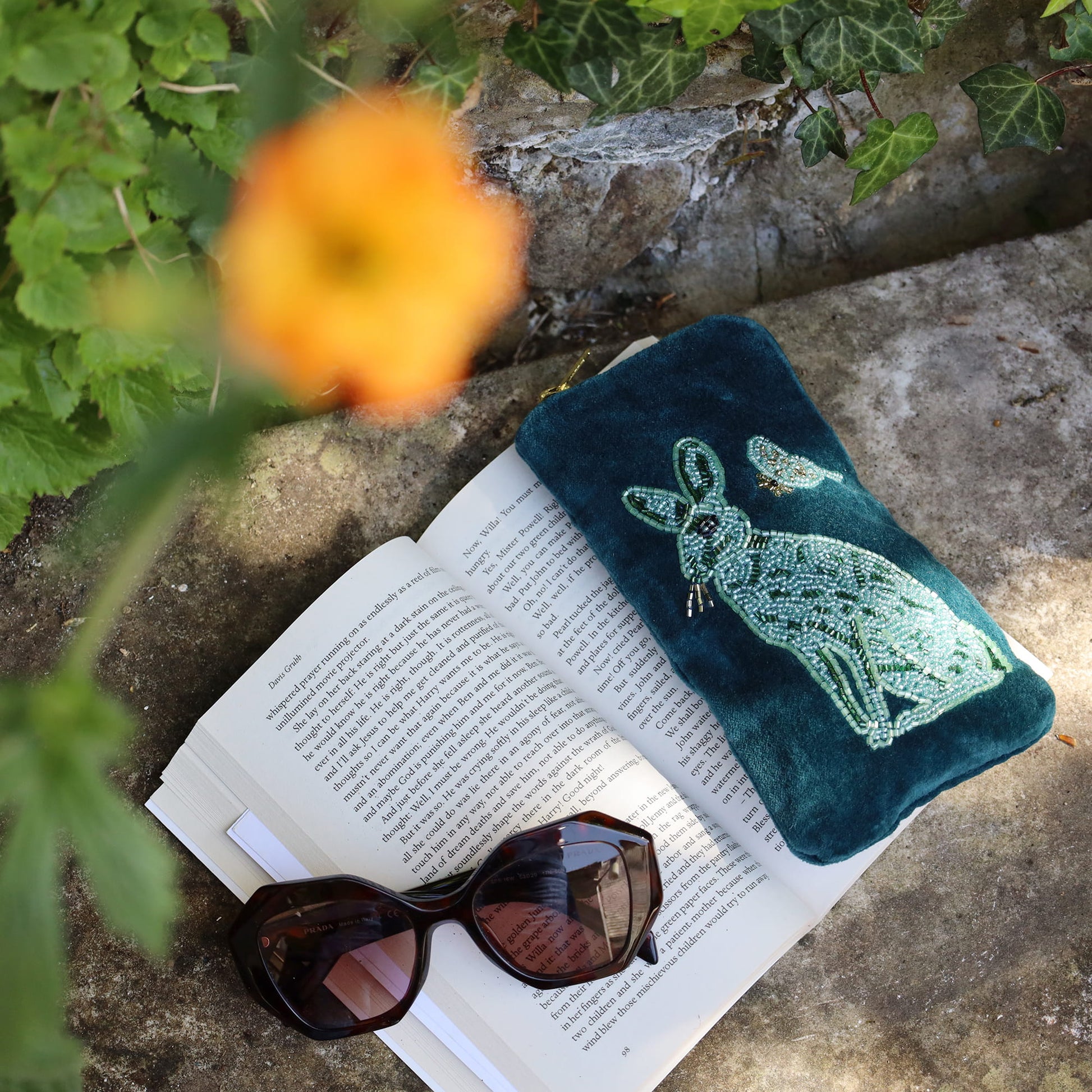 Green embroidered pouch with a rabbit design on an open book with sunglasses, surrounded by plants.