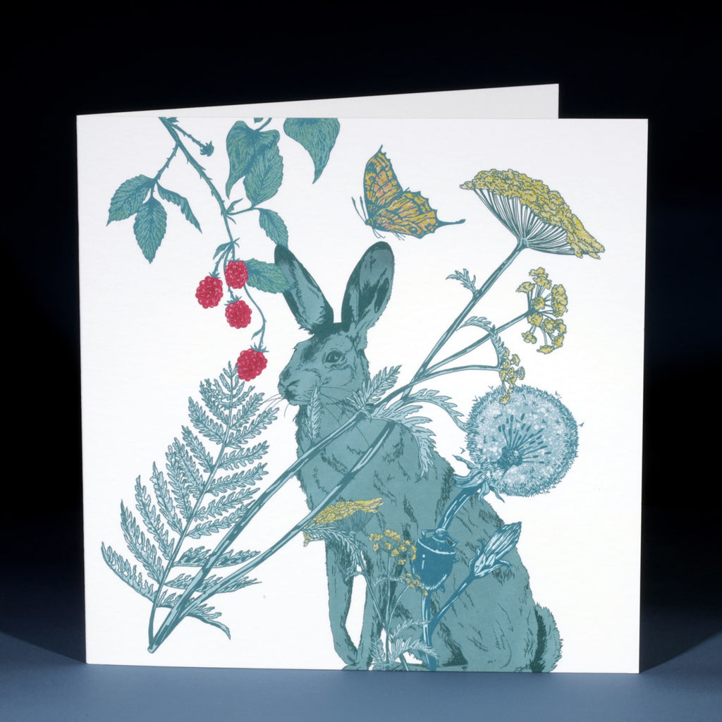 A square greeting card featuring a beautifully detailed illustration of a hare surrounded by botanical elements. The hare is drawn in muted green tones and is framed by delicate plants, including ferns, dandelions, cow parsley, raspberries, and a yellow butterfly. The illustration has a vintage, nature-inspired aesthetic on a crisp white background.