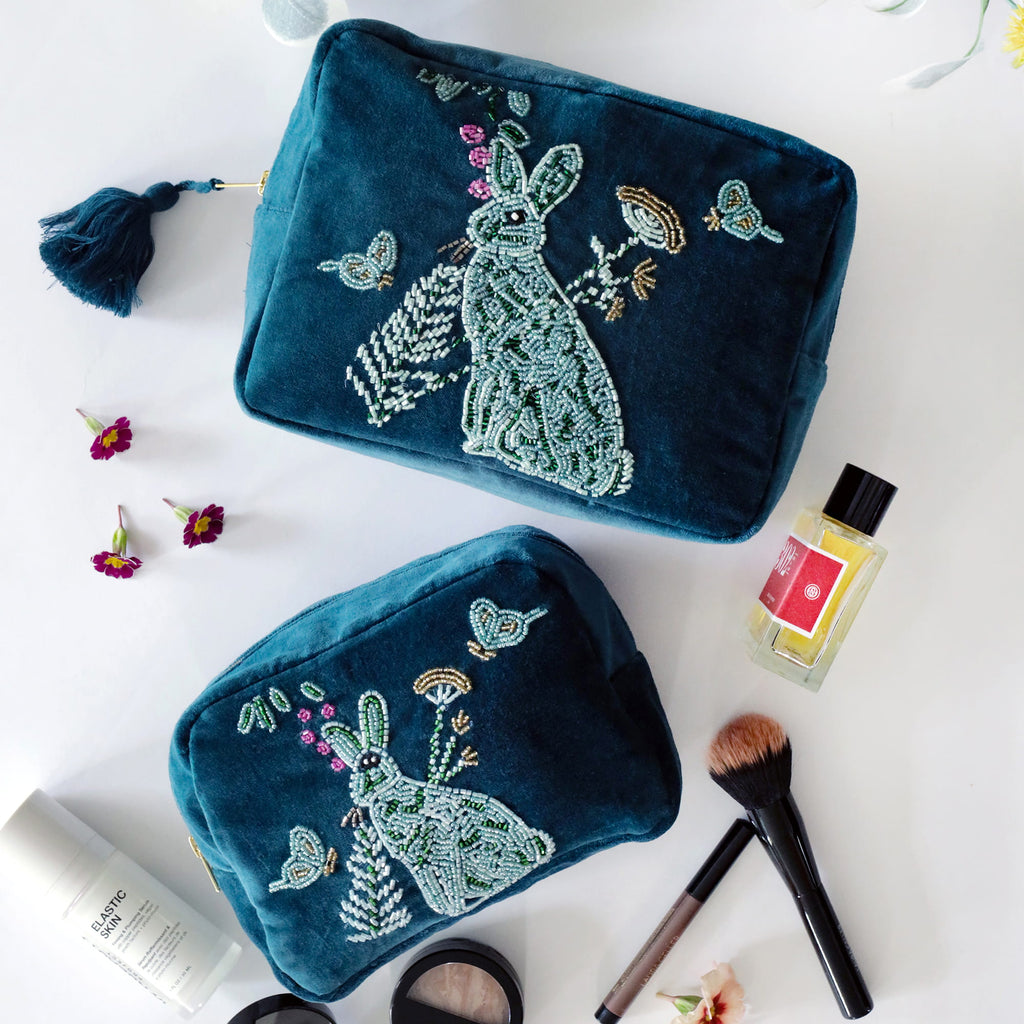 Two teal beaded pouches with hare designs on a white surface with makeup items.