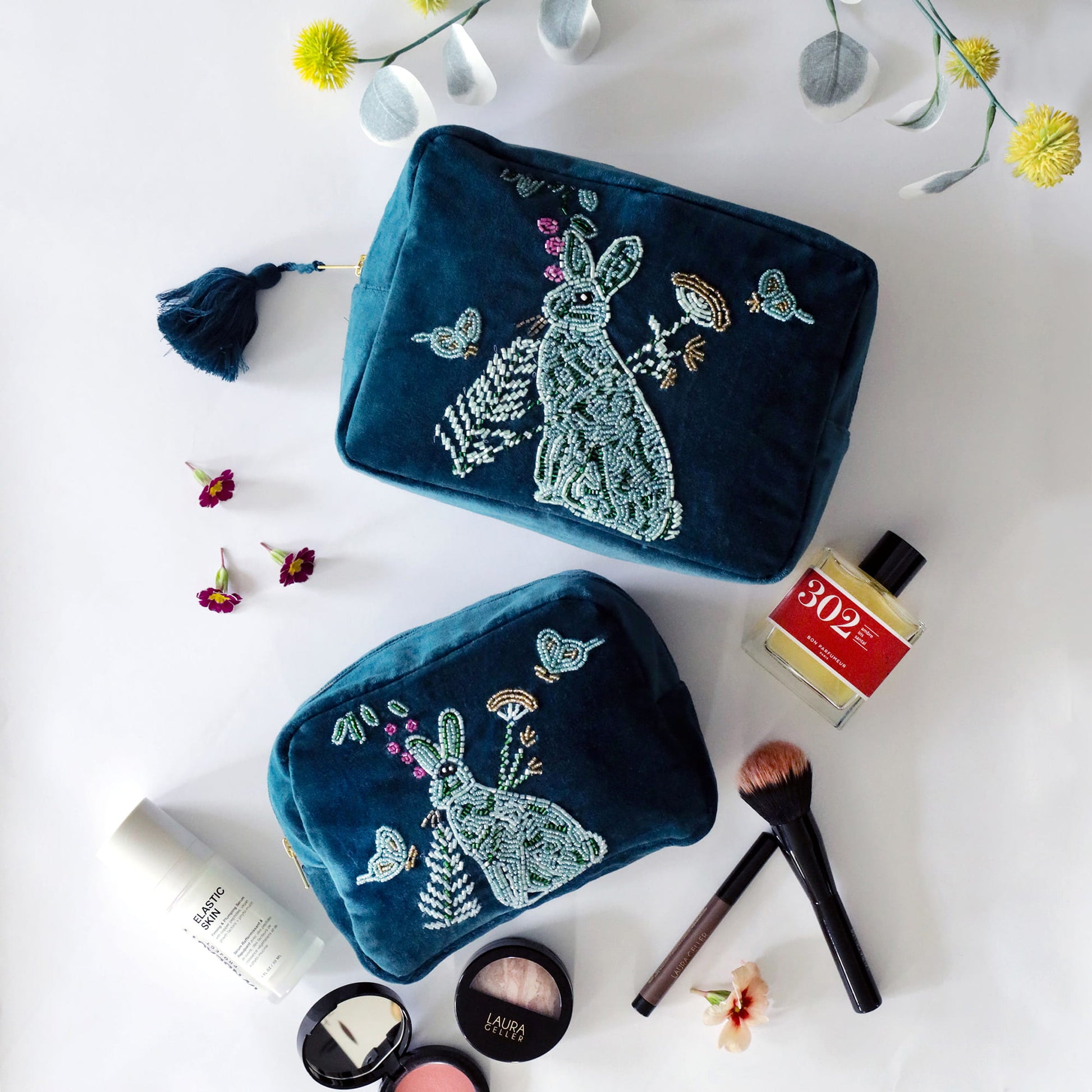 Two teal beaded pouches on a white surface with makeup items and flowers.