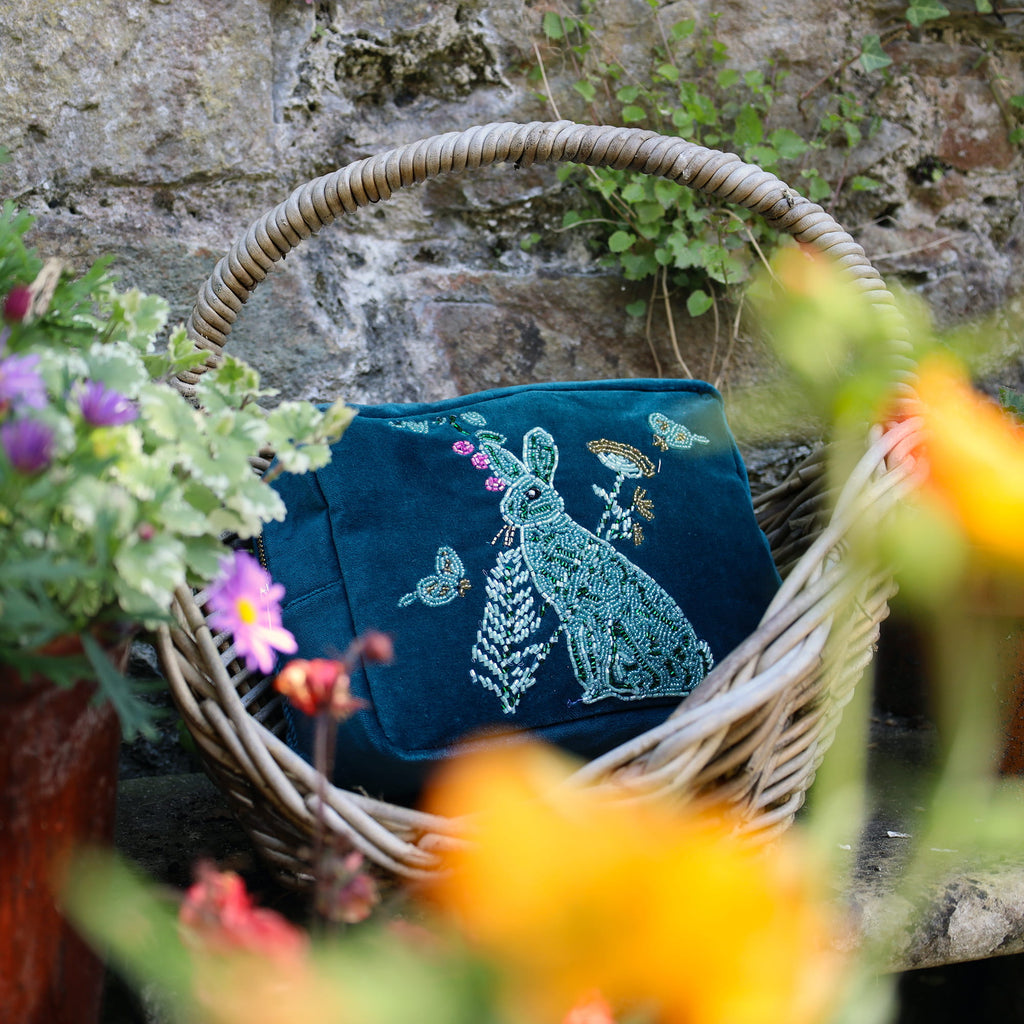 Embroidered blue bag with a rabbit design in a wicker basket surrounded by flowers and a stone wall.
