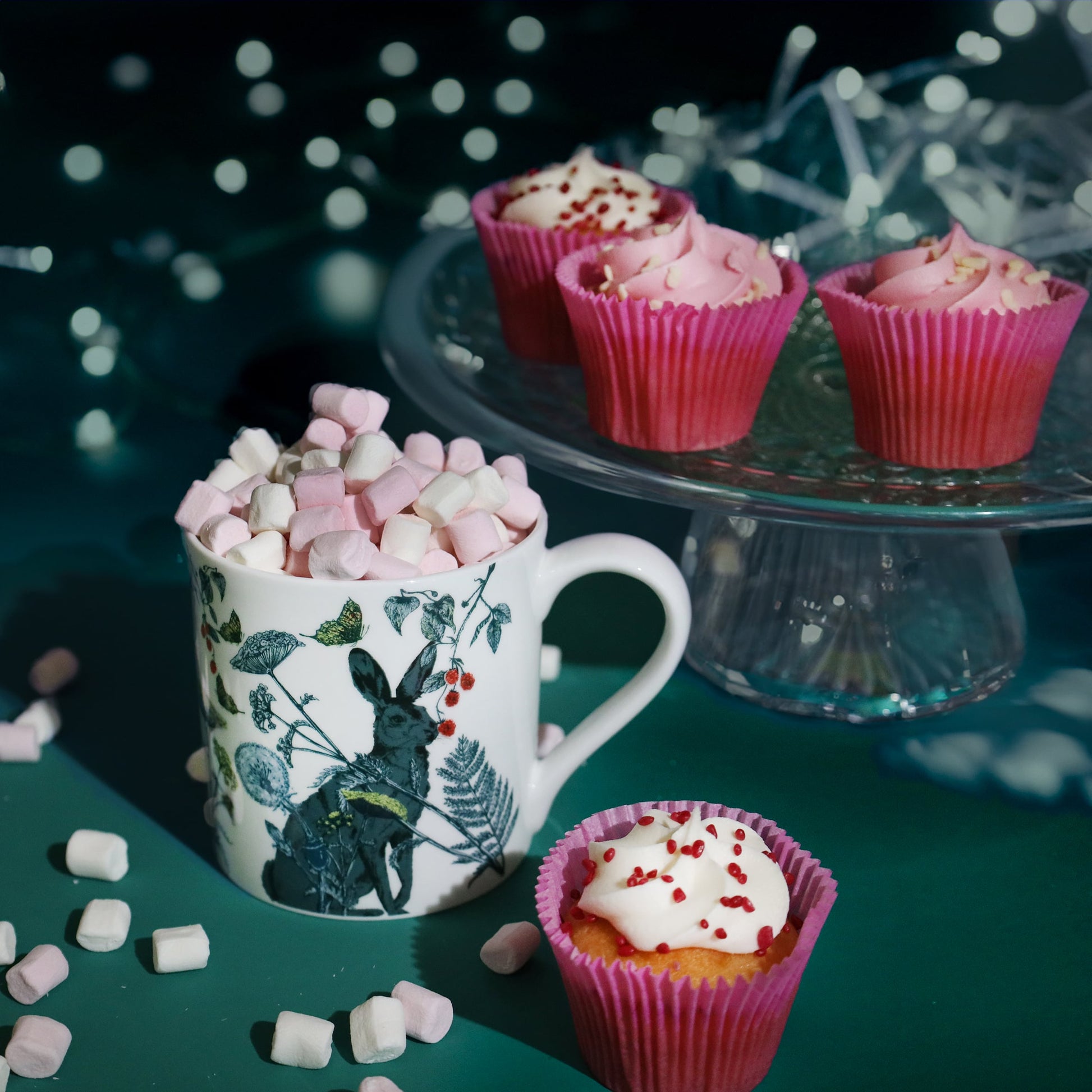 A festive scene featuring a white mug decorated with a hare and botanical design, filled with pink and white marshmallows, surrounded by scattered marshmallows and pink cupcakes with white icing and red sprinkles, against a dark teal background with soft glowing fairy lights.