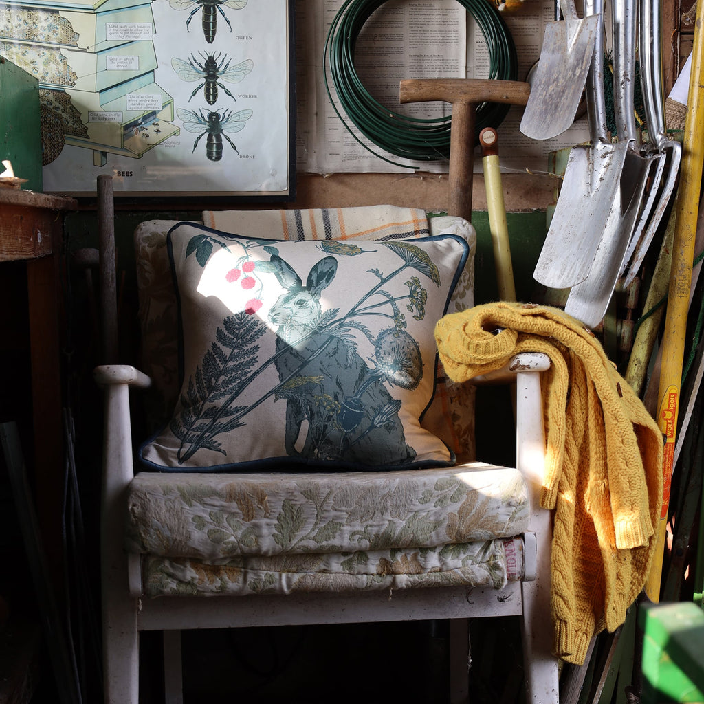 Rustic garden shed with a vintage white wooden chair, layered with a floral cushion and a hare-themed pillow featuring botanical details. A mustard-yellow knit sweater drapes over the armrest. Surrounding tools, including shovels, a spade, and coiled wire, hang on the walls. A bee illustration chart adds a nostalgic touch to the cozy, well-used space.