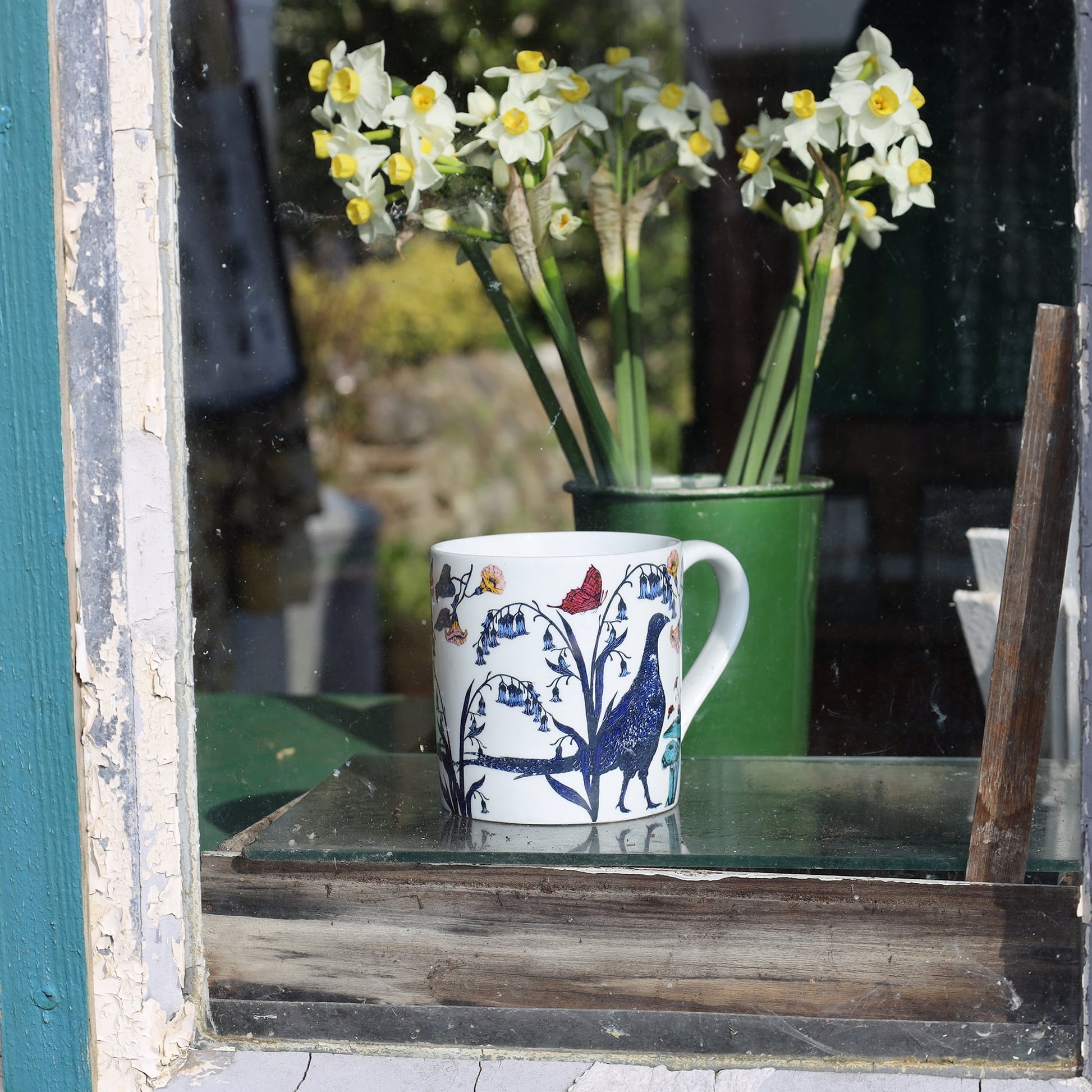 Bone china mug with a pheasant and wildflower design sitting on a windowsill, with a green jug of daffodils in the background.