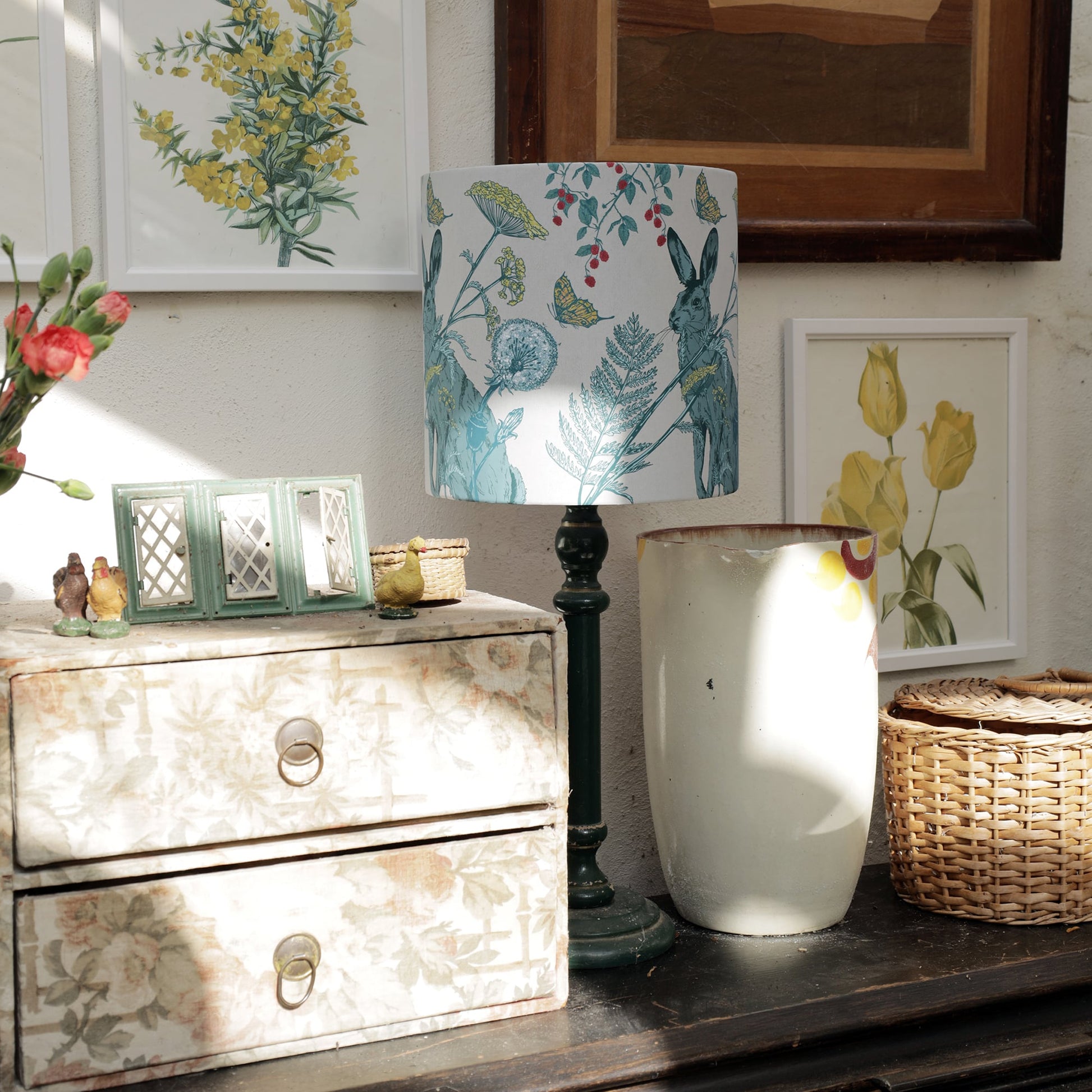 Sunlit corner featuring a botanical rabbit-print lampshade on a dark green lamp base, surrounded by vintage floral artwork, a weathered wooden drawer unit, and woven baskets. Soft natural light casts shadows, adding warmth to the rustic decor.