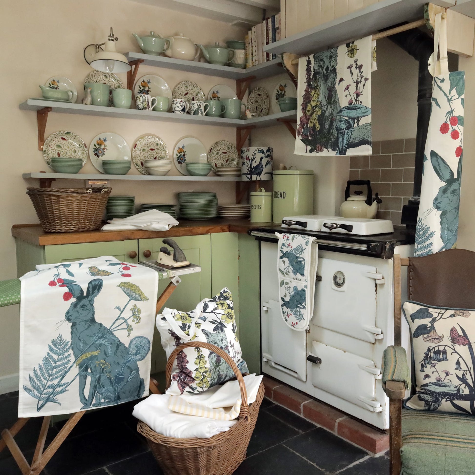 Cozy country kitchen with vintage green cabinets, open shelves displaying floral and pastel dishware, and a classic white Aga stove. Nature-inspired textiles, including hare and botanical print tea towels, tote bags, and cushions, add charm. A wicker basket with linens and an ironing board with a hare-print cloth enhance the rustic feel.