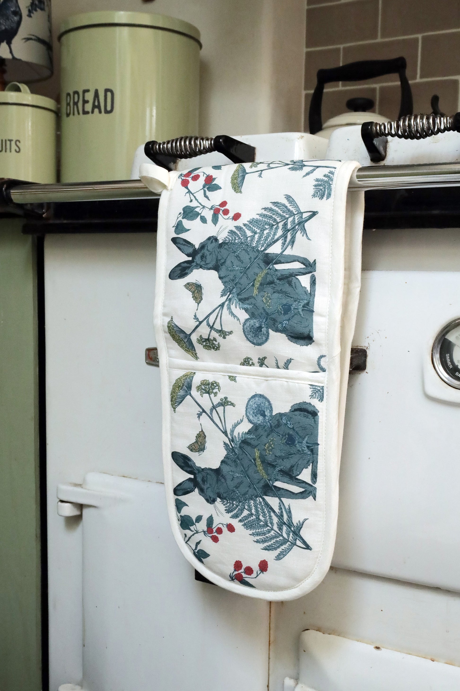 Close-up of a white Aga stove with a pair of hare-themed oven gloves draped over the handle. The gloves feature a detailed botanical illustration of a hare, ferns, berries, and butterflies. In the background, pastel green bread and biscuit tins sit on a wooden countertop, adding to the vintage country kitchen aesthetic