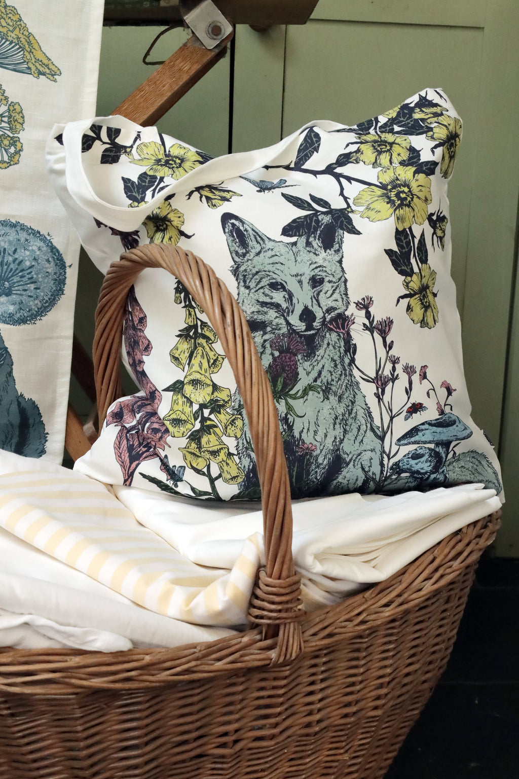 Close-up of a nature-inspired cushion featuring a fox surrounded by wildflowers, mushrooms, and insects, nestled in a wicker basket filled with folded linens in soft cream and yellow tones. The setting exudes a cozy, rustic charm with muted green cabinetry in the background.