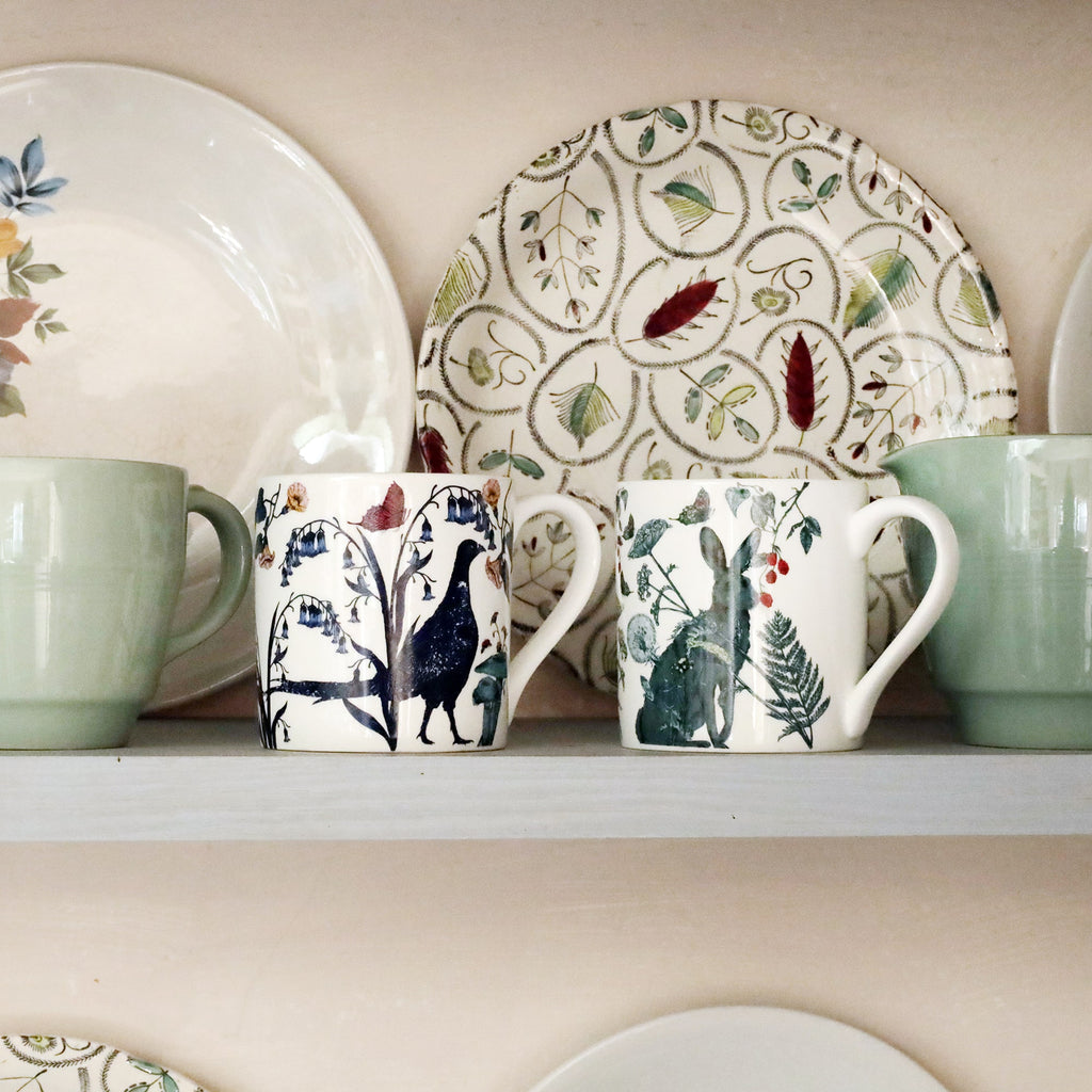 Two illustrated bone china mugs featuring a pheasant and a hare, displayed on a shelf with pastel green mugs and vintage floral-patterned plates in the background.