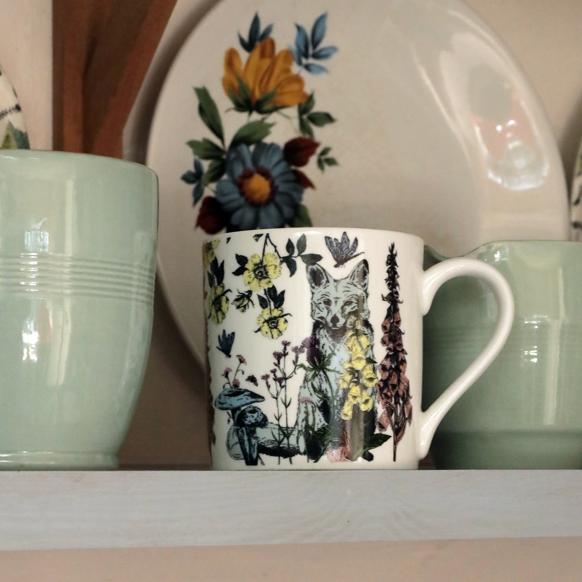 Bone china mug featuring a fox and wildflower illustration displayed on a shelf between two pale green mugs, with a floral vintage plate in the background.