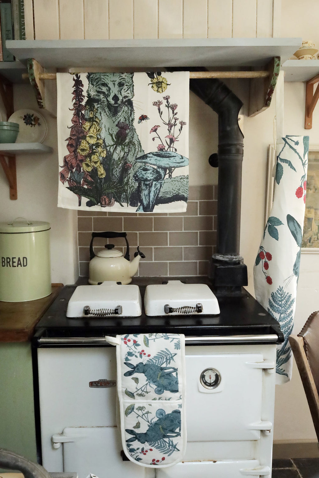 Vintage white Aga stove in a cozy country kitchen, adorned with nature-inspired textiles. A tea towel featuring a fox among wildflowers hangs above, while hare-themed oven gloves drape over the handle. A cream kettle sits on the stovetop, with pastel green bread and biscuit tins on the wooden countertop, adding to the rustic charm.