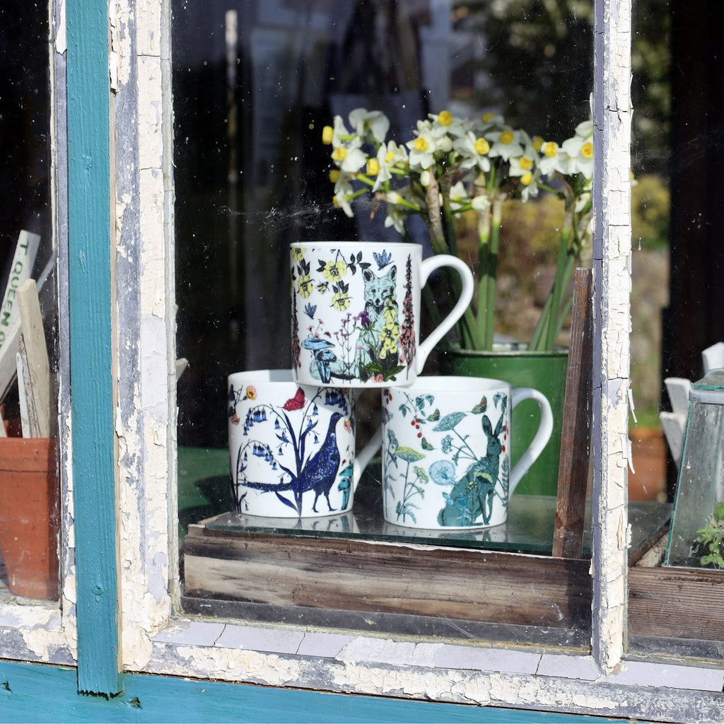 Three illustrated bone china mugs featuring wildlife and floral designs displayed on a rustic windowsill, with daffodils and garden tools in the background.