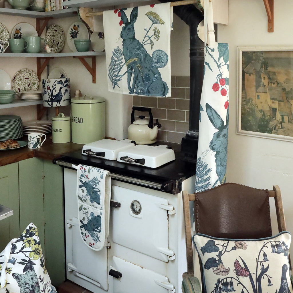 Cozy vintage-style kitchen featuring an Aga stove with decorative items from Cream Cornwall's Hedgerow collection, including a tea towel, apron, and oven gloves adorned with an illustrated hare and botanical elements. A white kettle sits on the stove, surrounded by pastel green tins labeled “BREAD” and “BISCUITS,” ceramic mugs and plates, and a cushion with floral and wildlife prints on a wooden chair nearby.