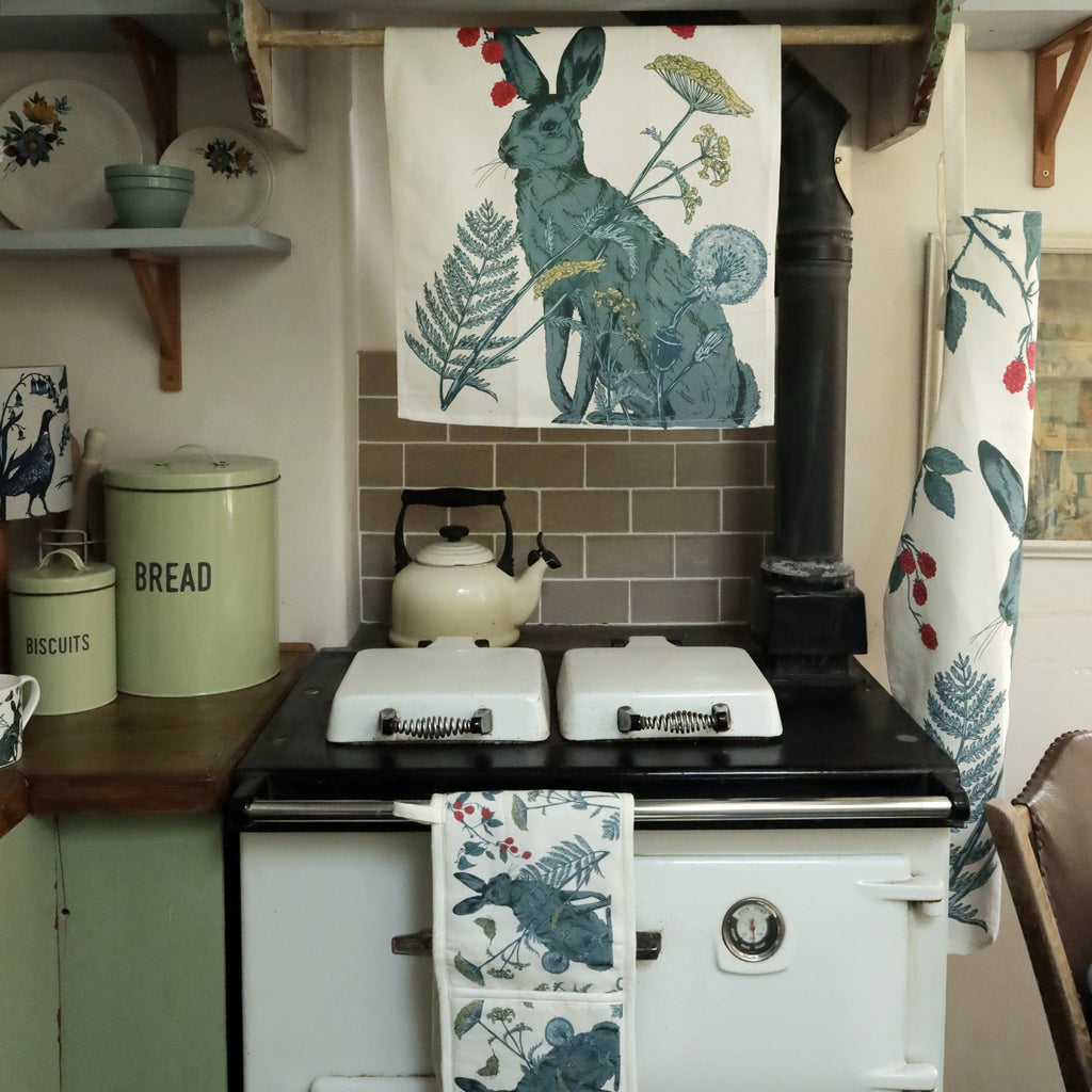 Vintage country kitchen with a classic white Aga stove, a cream kettle, and pastel green biscuit and bread tins. A hare-themed tea towel hangs above the stove, with matching nature-inspired textiles on the oven rail and side. Open shelves display floral dishware, adding to the cozy rustic charm.