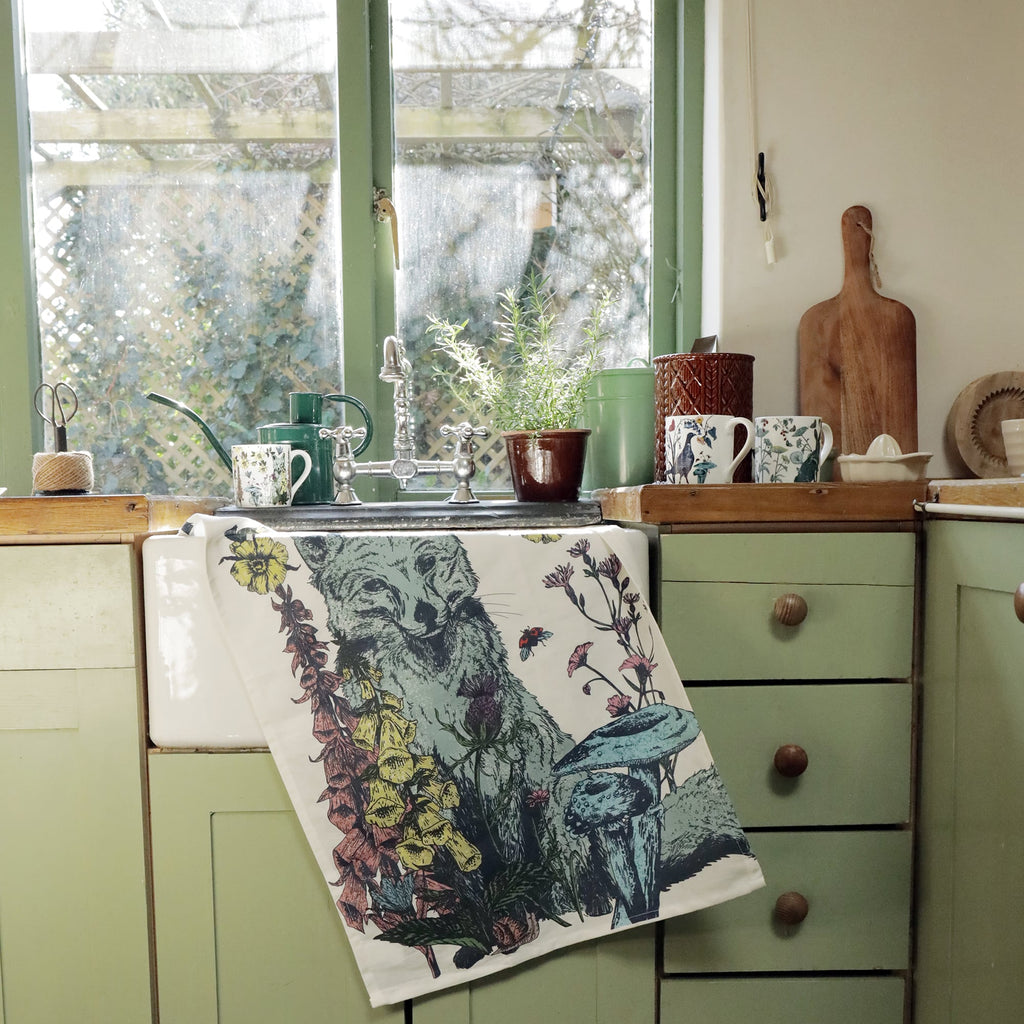 Rustic country kitchen with sage green cabinets and a farmhouse sink, draped with a tea towel featuring a fox surrounded by wildflowers and mushrooms. A large window lets in natural light, overlooking a garden. The wooden countertop holds potted herbs, floral mugs, a watering can, and kitchen essentials, creating a cozy, vintage-inspired setting.