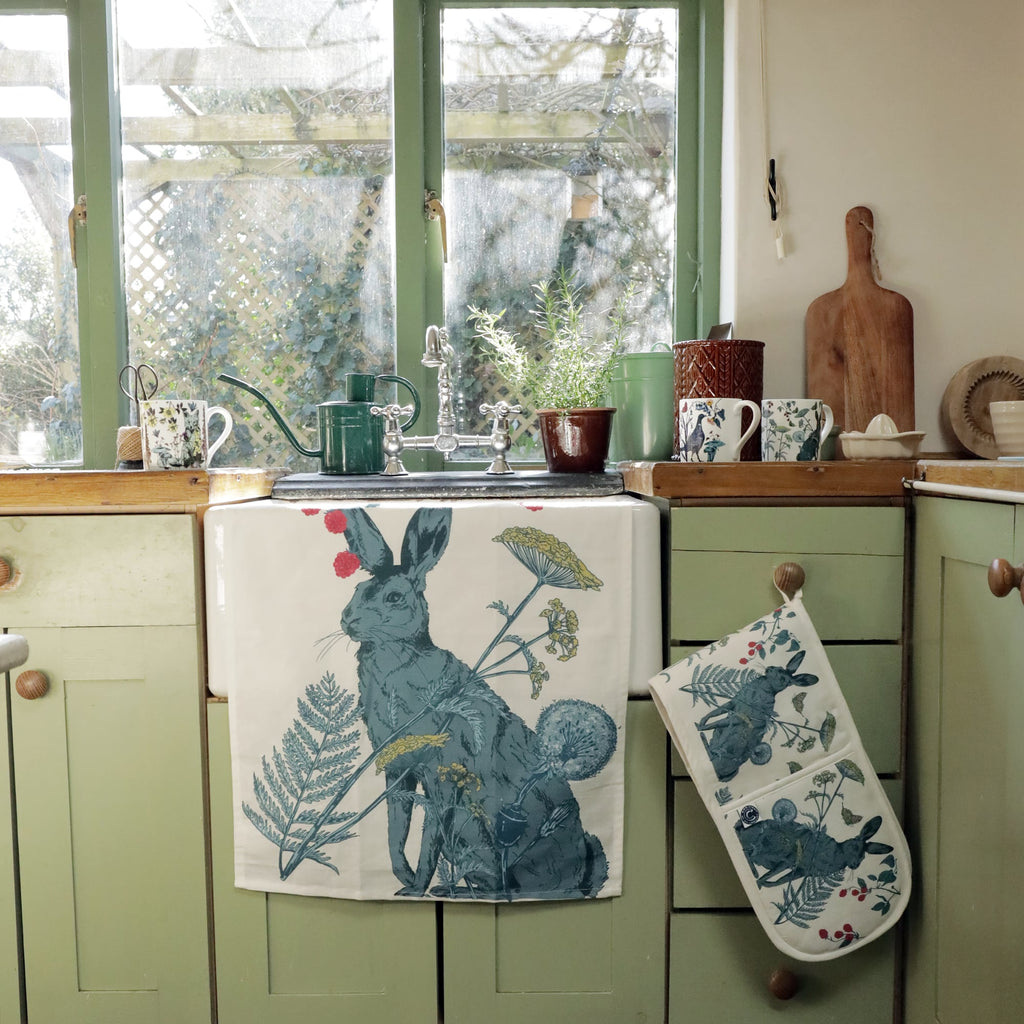 Rustic country kitchen with sage green cabinets and a farmhouse sink, set against a large window overlooking a garden. A hare-themed tea towel hangs over the sink, with matching oven gloves on a cabinet handle. The wooden countertop holds mugs, potted herbs, a watering can, and kitchen essentials, adding to the cozy, vintage charm.
