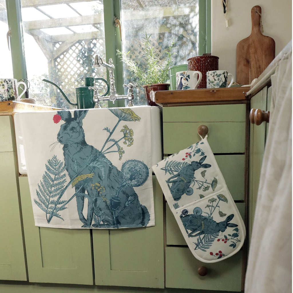 Cozy country kitchen with sage green cabinets and a farmhouse sink, featuring a hare-themed tea towel draped over the front and matching oven gloves hanging from a wooden knob. The countertop holds floral mugs, potted herbs, a green watering can, and a wooden chopping board, with a large window overlooking a sunlit garden.