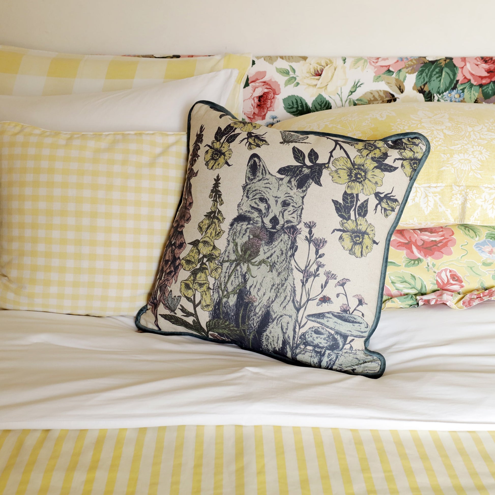 Cozy bedroom scene with a decorative cushion featuring a fox surrounded by wildflowers and foliage, resting on a bed with floral and gingham-patterned bedding in soft pastel tones.