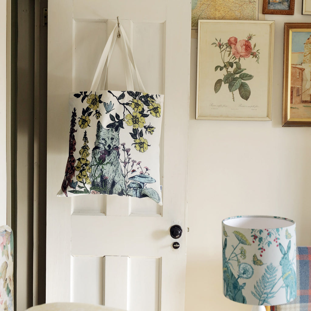 White wooden door with a nature-inspired tote bag hanging from a hook, featuring a detailed illustration of a fox surrounded by wildflowers and butterflies. The room has vintage charm with framed botanical and landscape prints on the wall. A matching floral-patterned lampshade in the foreground complements the soft, rustic aesthetic.