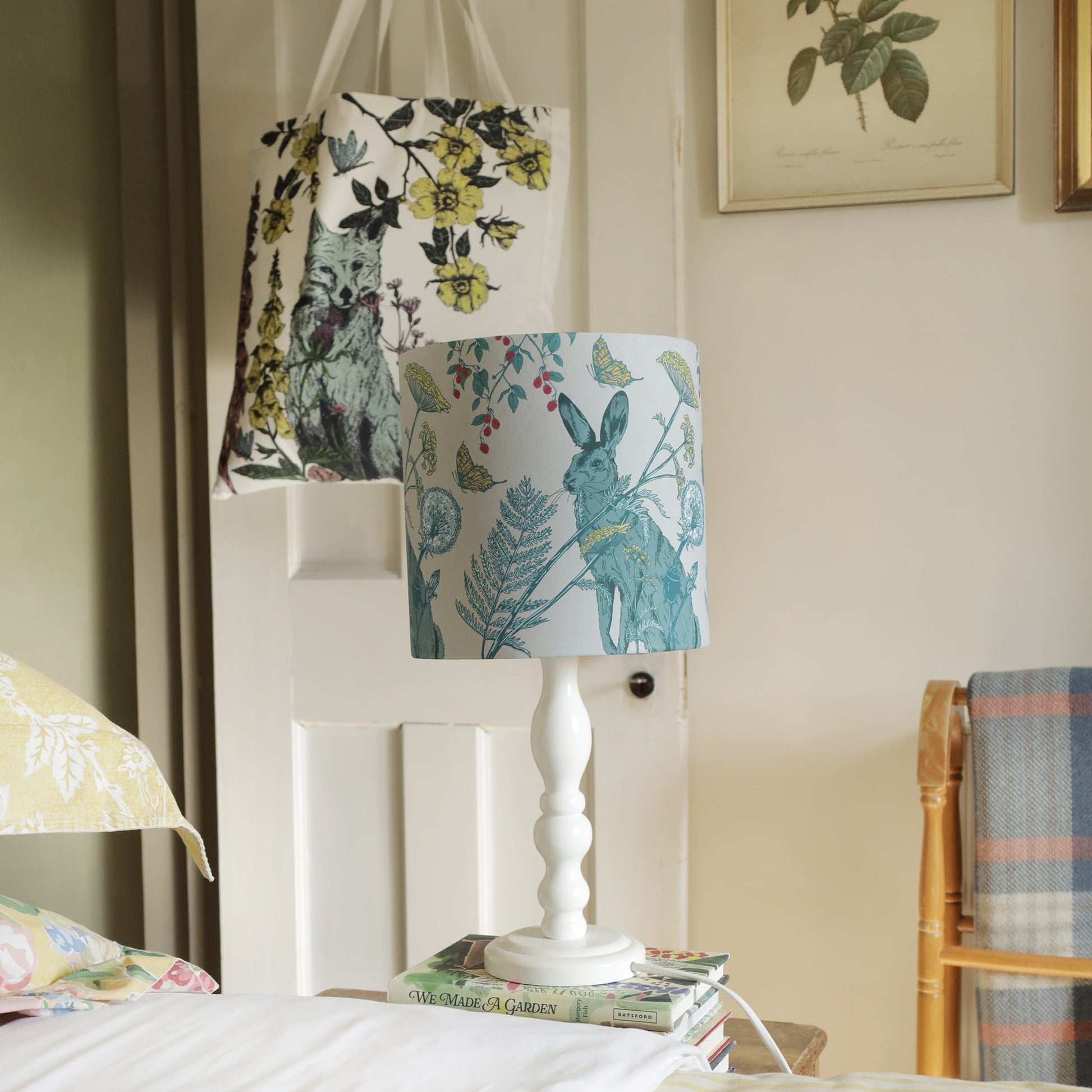 Cozy bedroom scene featuring a rabbit-themed lampshade on a white bedside lamp, resting on stacked garden books. A floral fox-print tote bag hangs on a wooden door, with vintage botanical artwork adorning the wall, creating a warm, rustic ambiance.