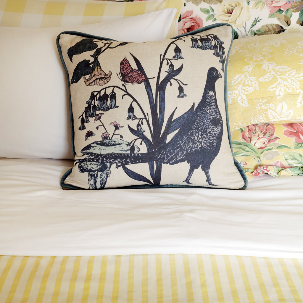 Cozy bed adorned with layered floral, gingham, and striped bedding in soft yellow and cream tones. A pheasant-themed cushion with botanical details sits at the center, adding a rustic countryside charm to the setting.