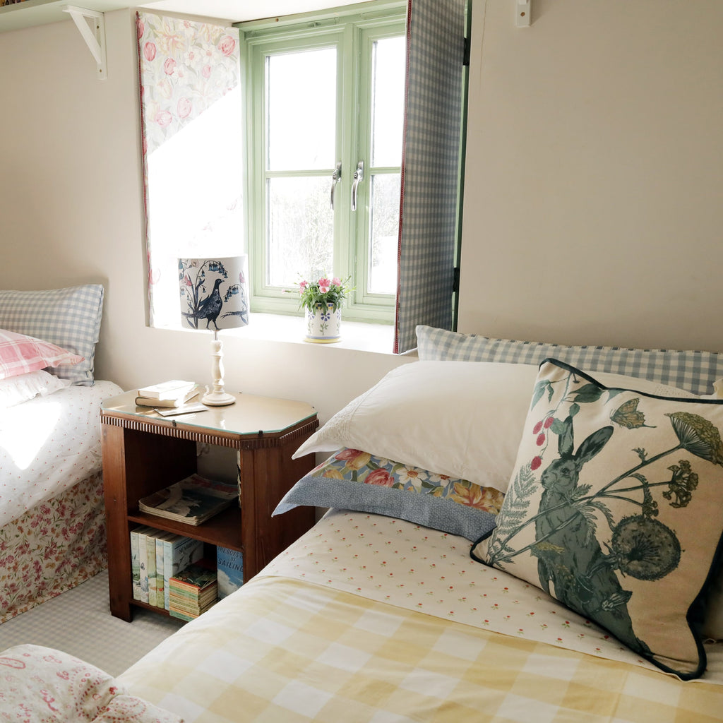 Cozy countryside bedroom with twin beds dressed in floral, gingham, and checkered bedding. A hare-themed cushion rests on one bed, complementing the soft pastel tones. A wooden bedside table with books and a bird-print lamp sits between the beds, while a green-paned window with floral shutters lets in natural sunlight.