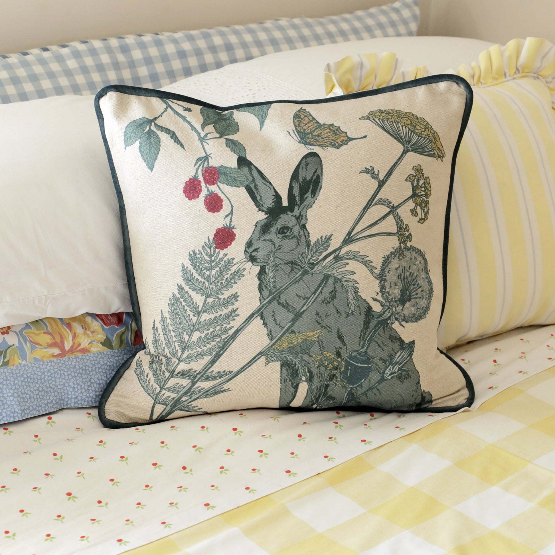 Close-up of a cozy bed adorned with a hare-themed cushion featuring a detailed botanical design of a hare, ferns, berries, and butterflies. The cushion sits among layered bedding with gingham, floral, and striped patterns in soft pastel shades of blue, yellow, and cream, creating a charming countryside aesthetic.