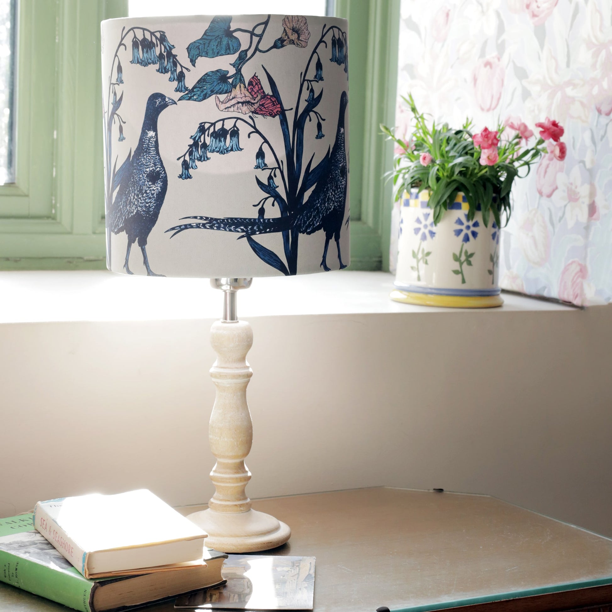 Cozy interior with a wooden table featuring a pheasant-themed lampshade adorned with botanical details. Sunlight streams through a green-paned window, illuminating a floral jug filled with pink flowers on the windowsill. A small stack of vintage books adds to the rustic charm of the space.