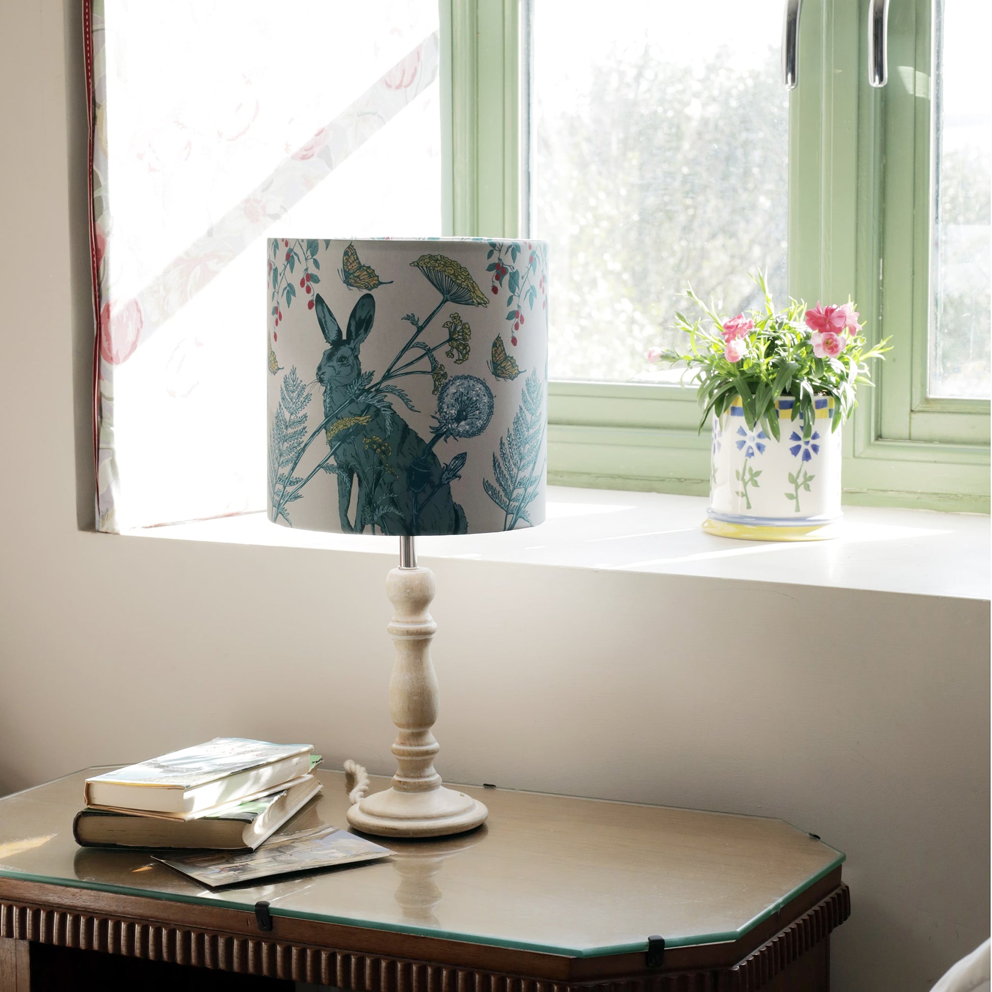 Sunlit bedside scene featuring a rabbit-themed lampshade on a white wooden lamp base, placed on a vintage glass-top nightstand. A stack of books and postcards rest beside it, while a floral ceramic pot with pink flowers sits on the windowsill, adding a charming country touch.