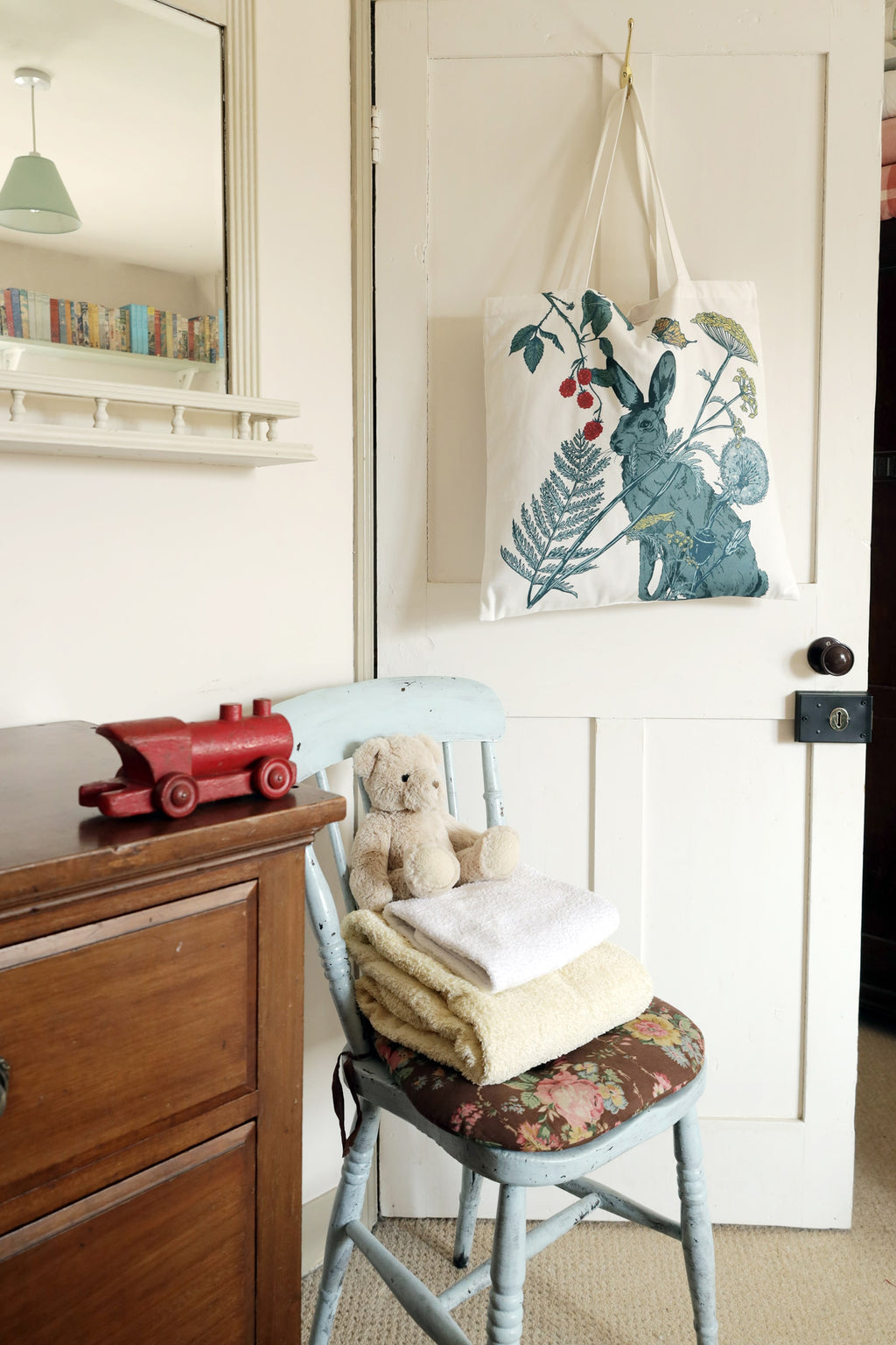 A cozy, vintage-inspired room with a white tote bag hanging on a wooden door. The tote bag features a nature-themed design with a blue hare, wildflowers, ferns, raspberries, and a butterfly. Below, a rustic pale blue wooden chair with a floral cushioned seat holds a neatly folded stack of towels and a plush teddy bear. A wooden dresser sits nearby with a red wooden toy train on top. A mirror above the dresser reflects bookshelves and a green pendant light, adding to the warm, nostalgic atmosphere.