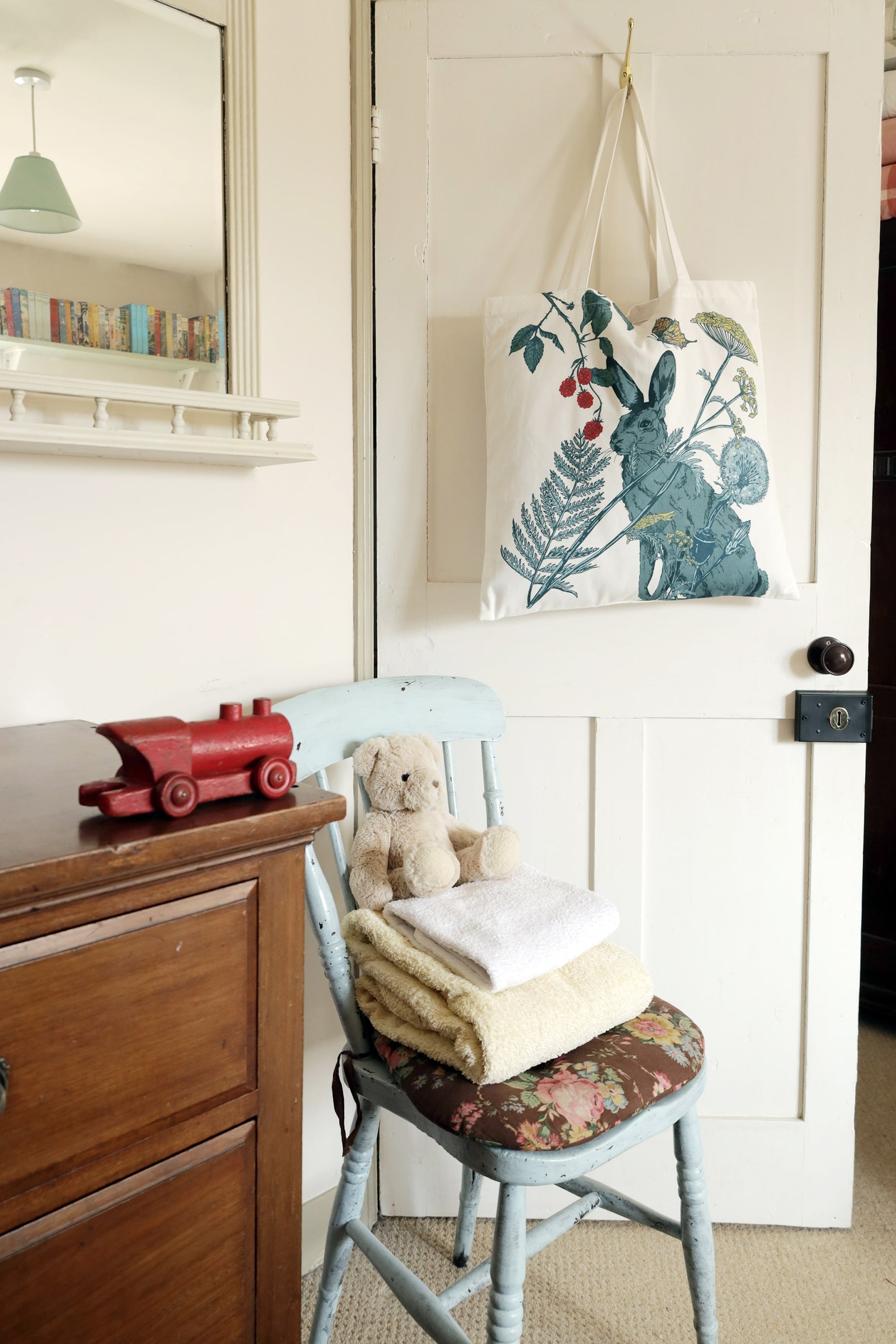 A cozy, vintage-inspired room with a white tote bag hanging on a wooden door. The tote bag features a nature-themed design with a blue hare, wildflowers, ferns, raspberries, and a butterfly. Below, a rustic pale blue wooden chair with a floral cushioned seat holds a neatly folded stack of towels and a plush teddy bear. A wooden dresser sits nearby with a red wooden toy train on top. A mirror above the dresser reflects bookshelves and a green pendant light, adding to the warm, nostalgic atmosphere.