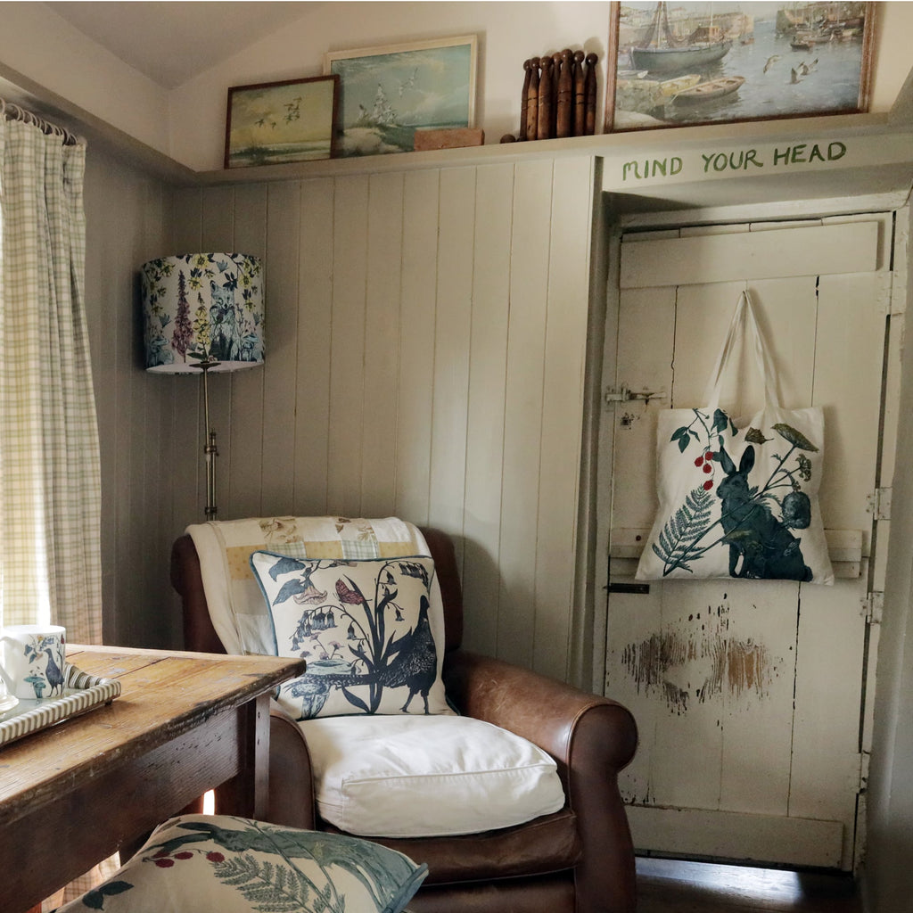 A cozy corner in a rustic cottage featuring a vintage leather armchair adorned with a nature-inspired cushion showcasing a pheasant and botanical illustrations. A matching tote bag hangs on a distressed wooden door, adding to the cottagecore charm. A tall floor lamp with a complementary woodland-patterned shade provides soft lighting. The wooden table beside the chair holds a cup and saucer, evoking a peaceful and inviting atmosphere