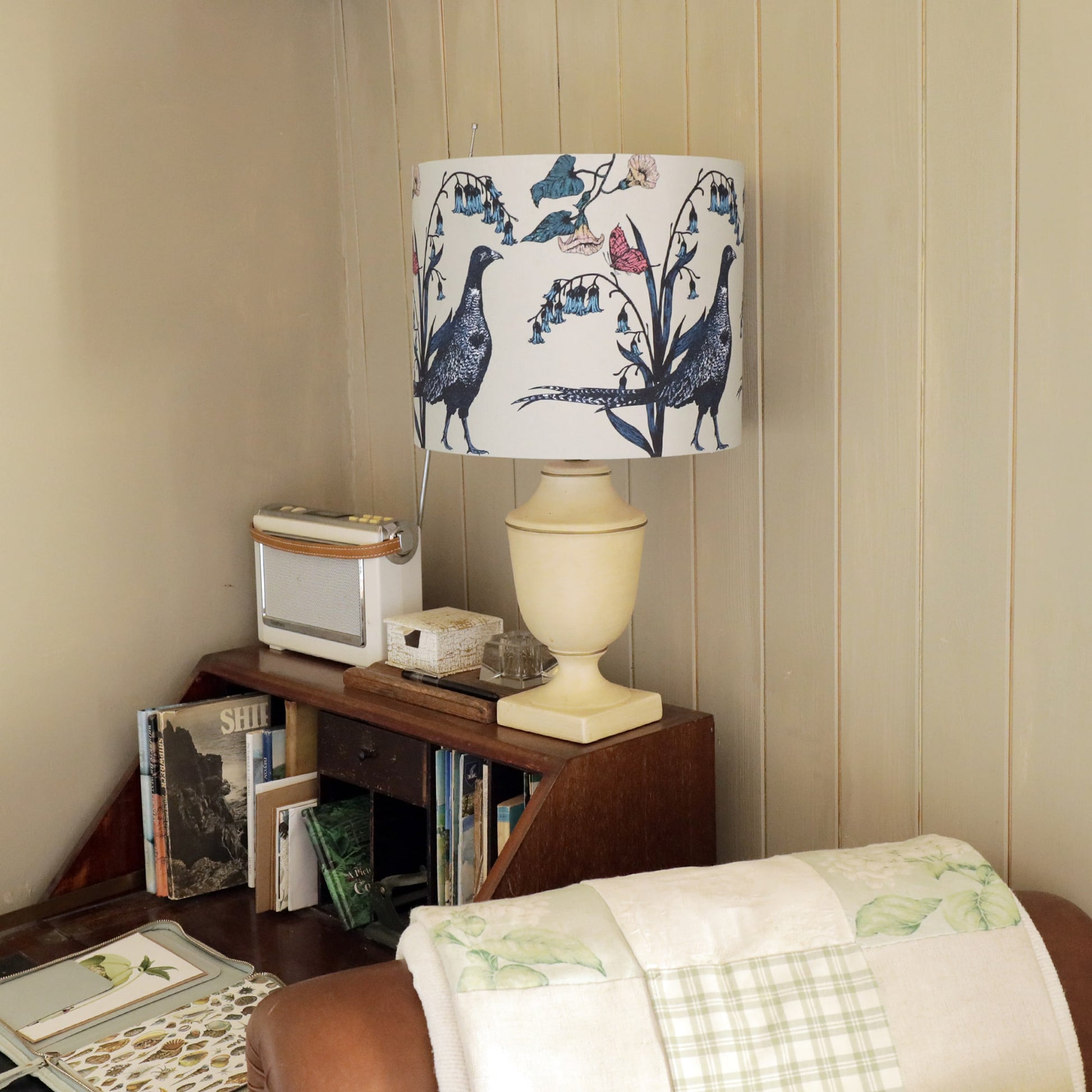 Cozy reading nook with a vintage wooden bookcase filled with books and decorative items. A cream-colored table lamp with a pheasant-themed botanical lampshade sits atop the bookcase, next to a retro radio. A quilted throw with soft green patterns drapes over a leather armchair, adding to the rustic charm of the space.