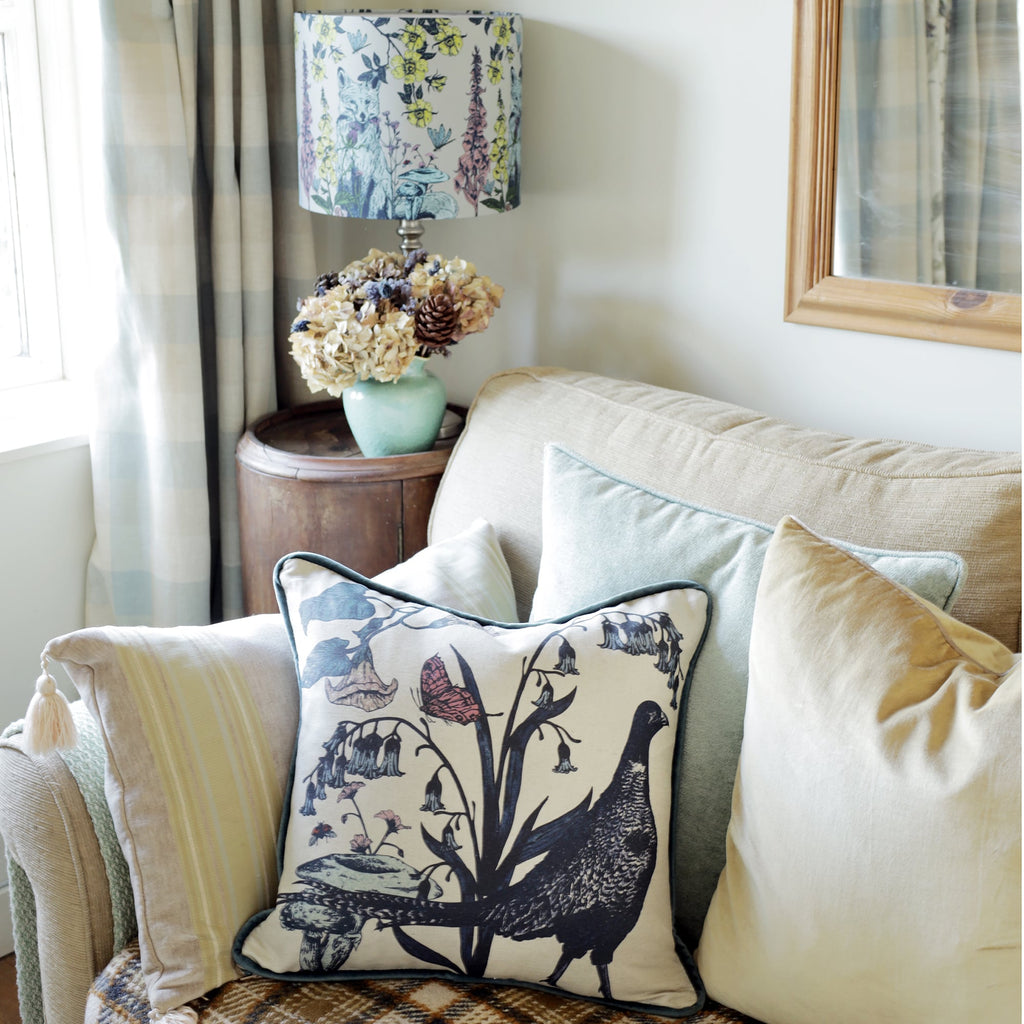 Cozy living space with a beige sofa adorned with layered cushions, including a pheasant-themed pillow featuring botanical details. A rustic wooden side table holds a vase of dried hydrangeas and a lamp with a floral-patterned shade. Soft pastel tones, plaid accents, and natural textures create a warm countryside charm.