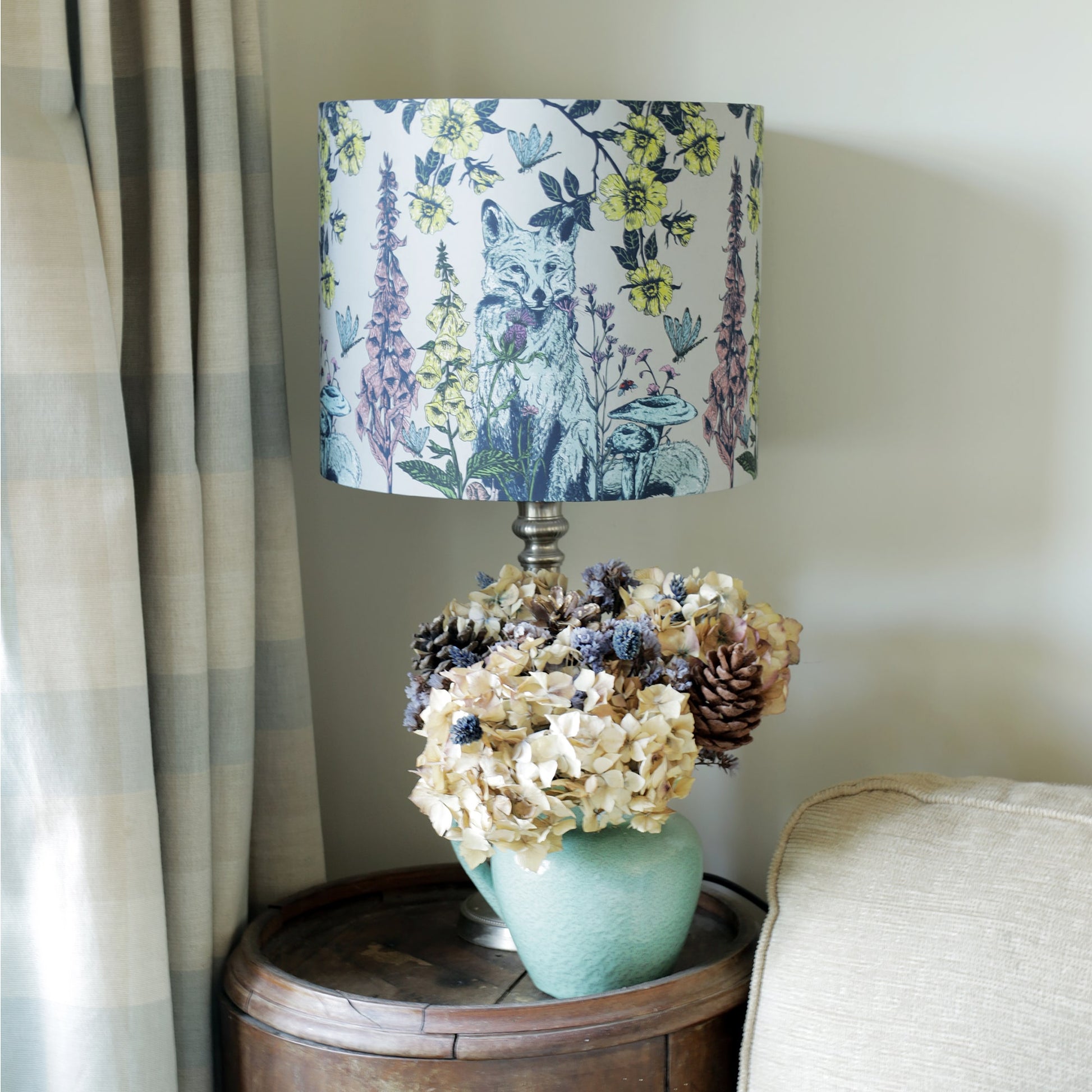 A cozy interior scene featuring a vintage-style table lamp with a nature-inspired lampshade adorned with illustrations of a fox, wildflowers, and butterflies. The lamp sits on a rustic wooden side table next to a ceramic vase filled with dried hydrangeas and pinecones, adding a touch of woodland charm. A soft, neutral-toned cushion rests against a sofa, complementing the serene and elegant decor.