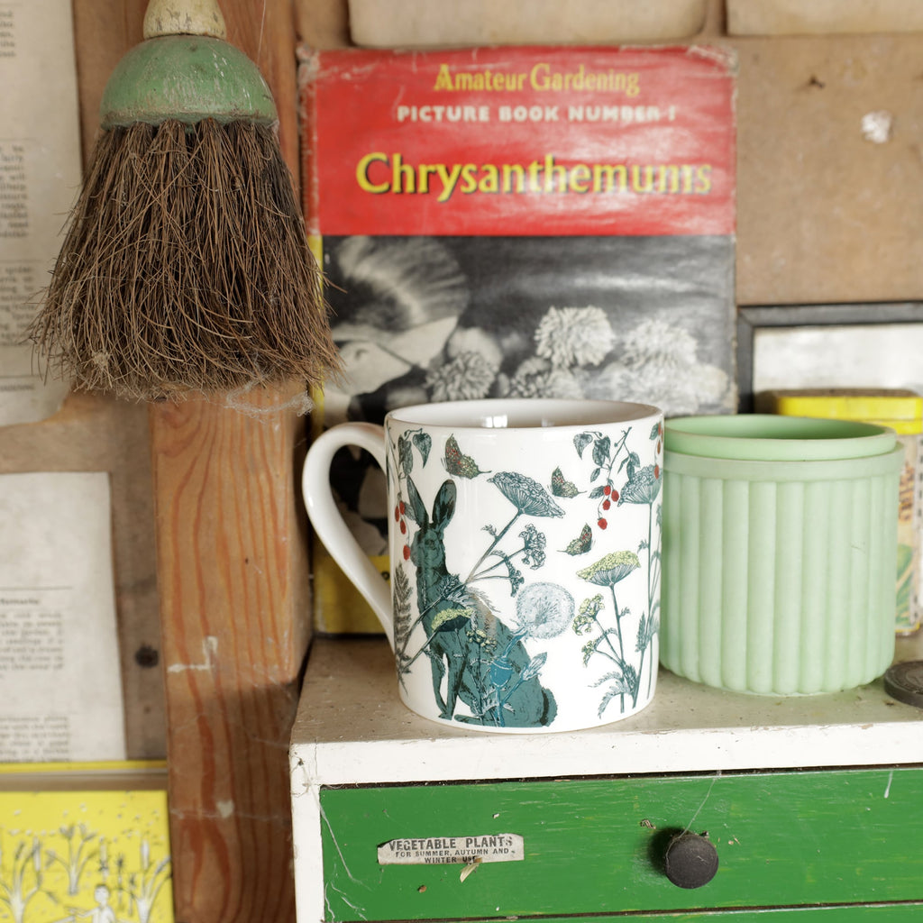 Bone china mug with a hare and botanical illustration displayed on a vintage green-and-white drawer unit, with a mint green pot and retro gardening book in the background.