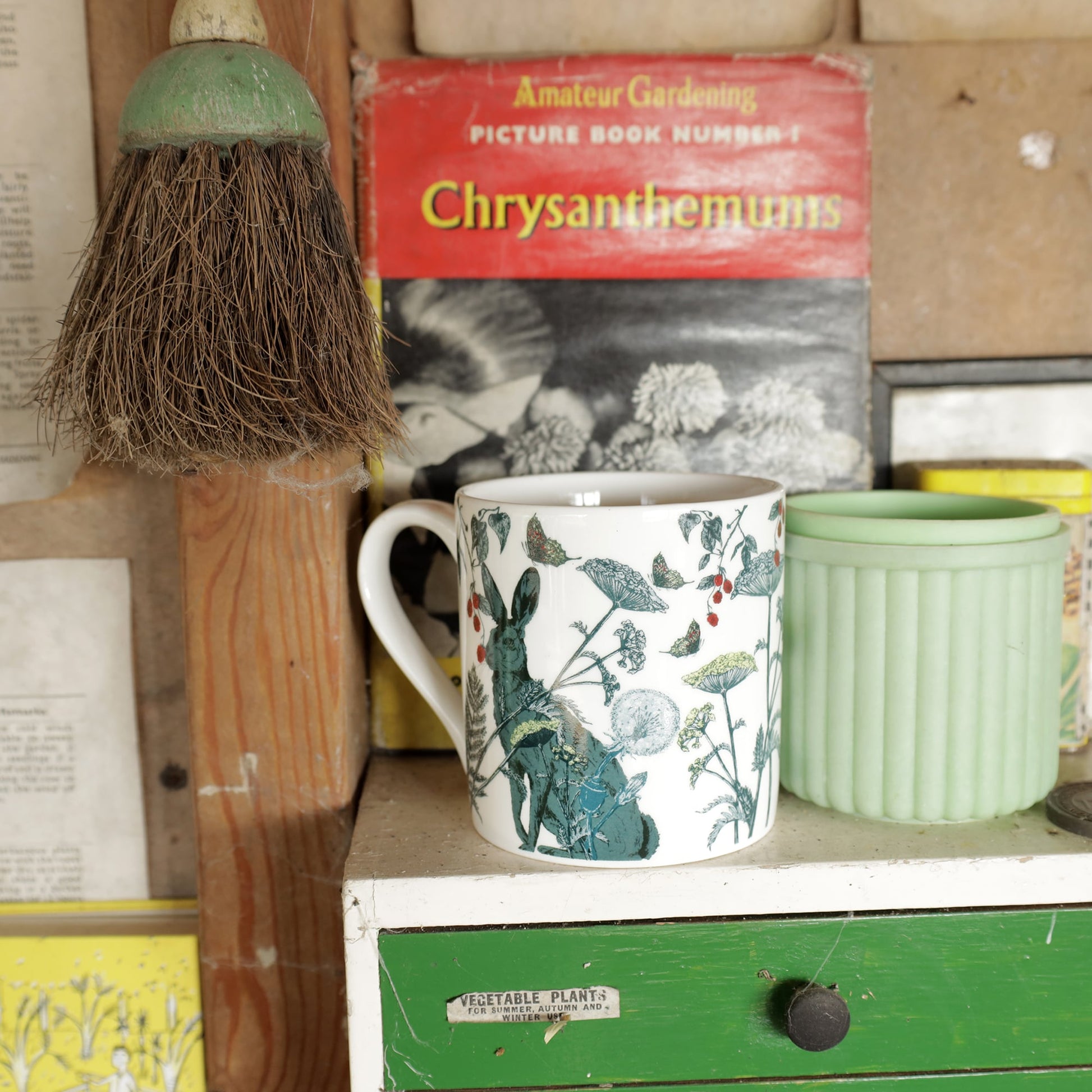 Bone china mug with a hare and botanical illustration displayed on a vintage green-and-white drawer unit, with a mint green pot and retro gardening book in the background.