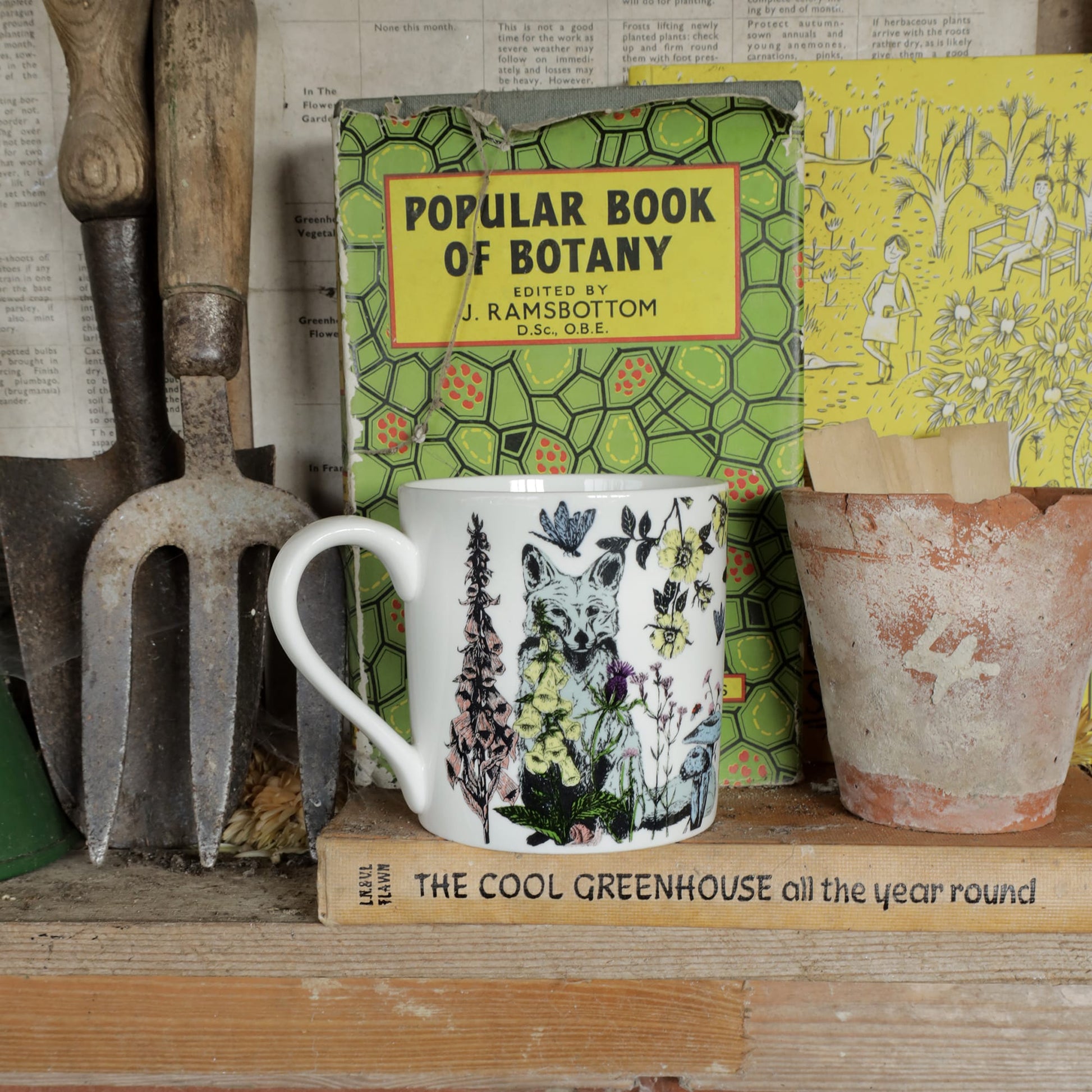 Illustrated bone china mug featuring a fox and wildflowers, displayed on a wooden shelf alongside vintage gardening tools, a terracotta pot, and botanical books.