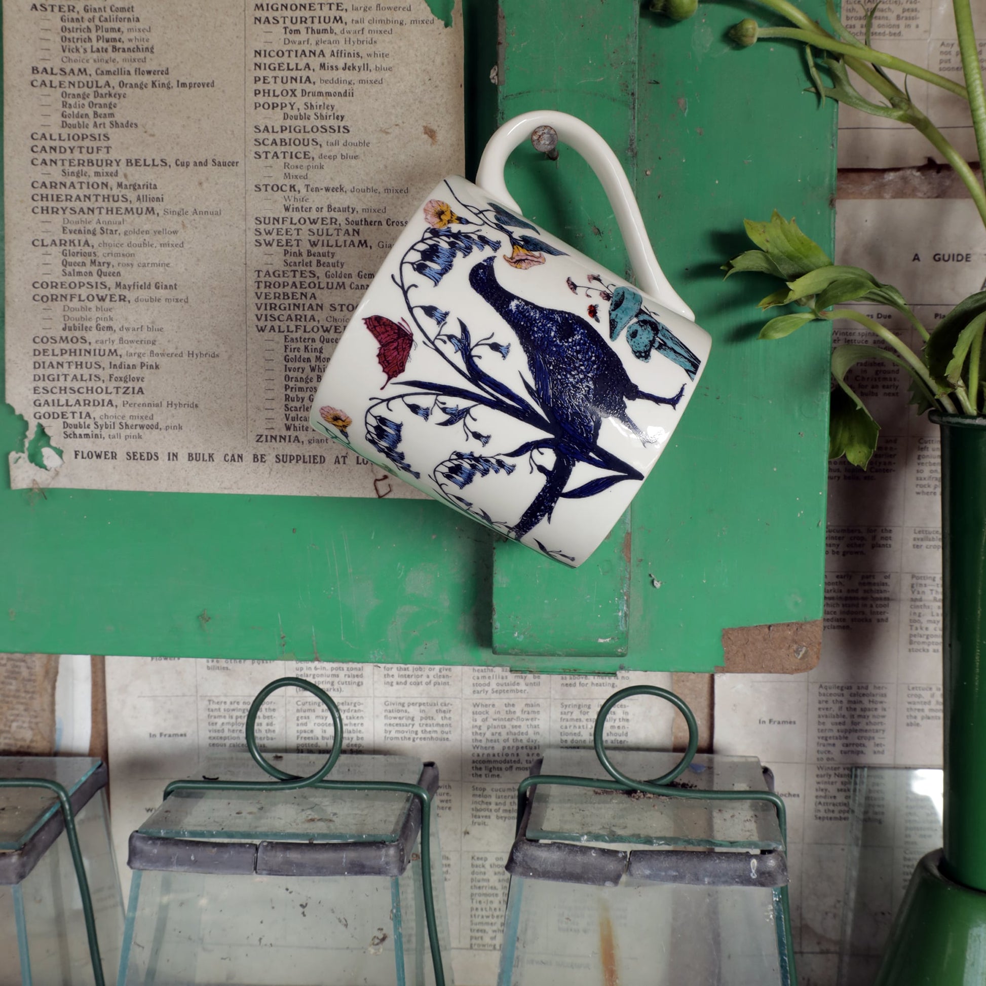 Bone china mug with a pheasant and wildflower illustration hanging on a green wooden shelf, with vintage seed catalog pages and glass cloches below.
