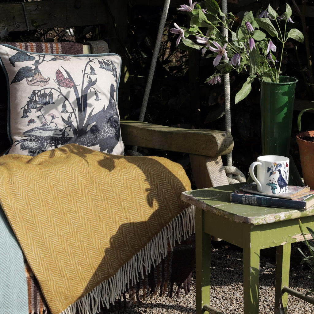 Cozy outdoor seating area with a wooden armchair draped in a mustard-yellow herringbone blanket and a pheasant-themed cushion. A weathered green side table holds a bird-print mug, books, and a vase of purple clematis flowers. Sunlight and shadows add warmth to this tranquil garden setting.