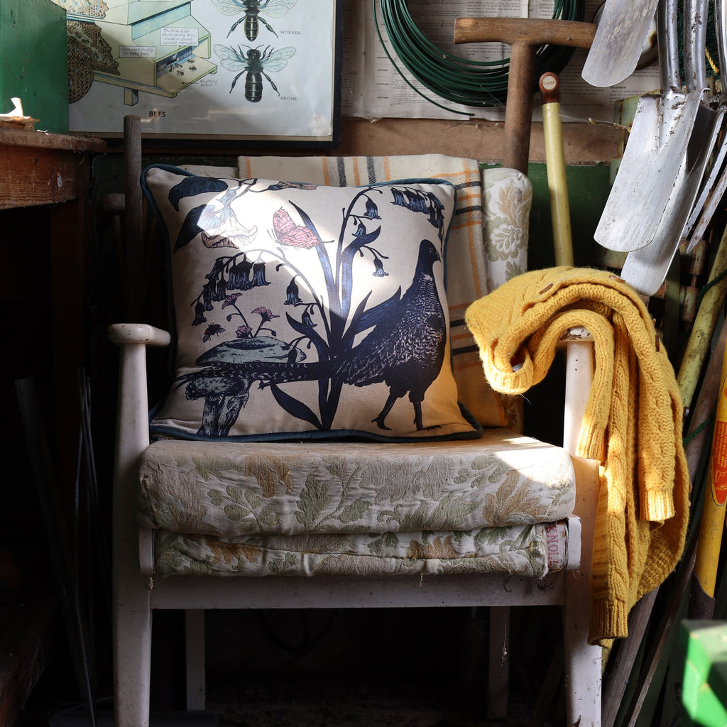 Rustic garden shed with a vintage white wooden chair, layered with a floral cushion and a pheasant-themed pillow featuring botanical details. A mustard-yellow knit sweater drapes over the armrest. Surrounding tools, including shovels, a spade, and coiled wire, hang on the walls. A bee illustration chart adds a nostalgic touch to the cozy, well-used space.
