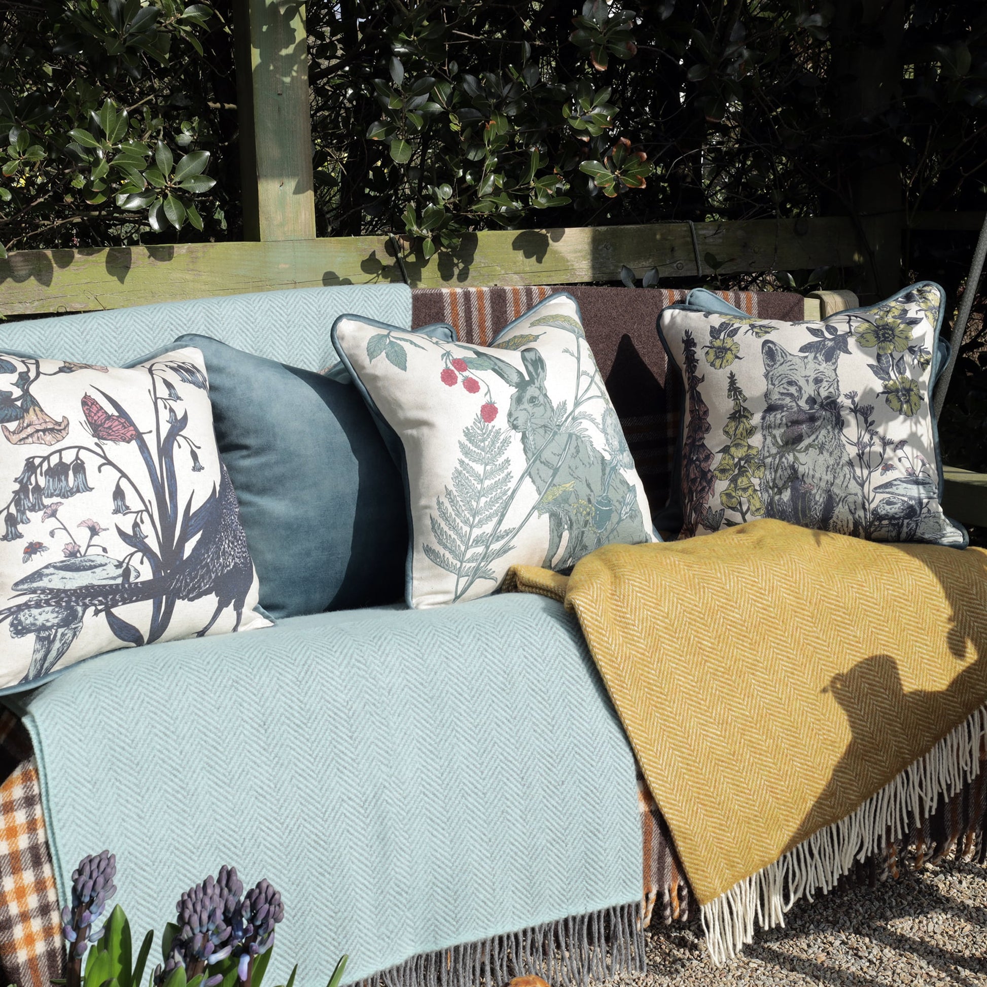 An inviting outdoor seating area featuring a wooden bench adorned with soft blankets and decorative cushions. The cushions showcase intricate wildlife and botanical illustrations, including a pheasant, a hare, and a fox among foliage. A mustard yellow throw adds warmth, while potted hyacinths in the foreground enhance the natural setting.