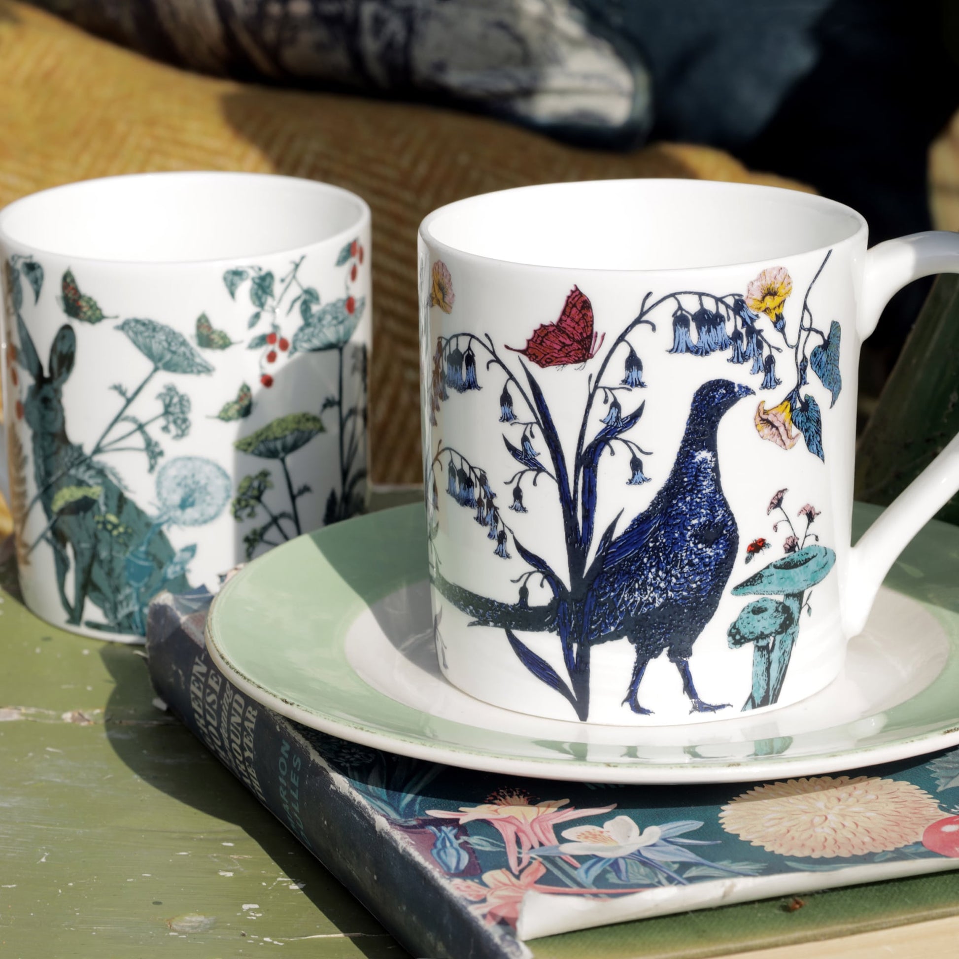 Two illustrated bone china mugs featuring countryside wildlife designs, including a pheasant and a hare, displayed on a green table with a vintage book and plate underneath.