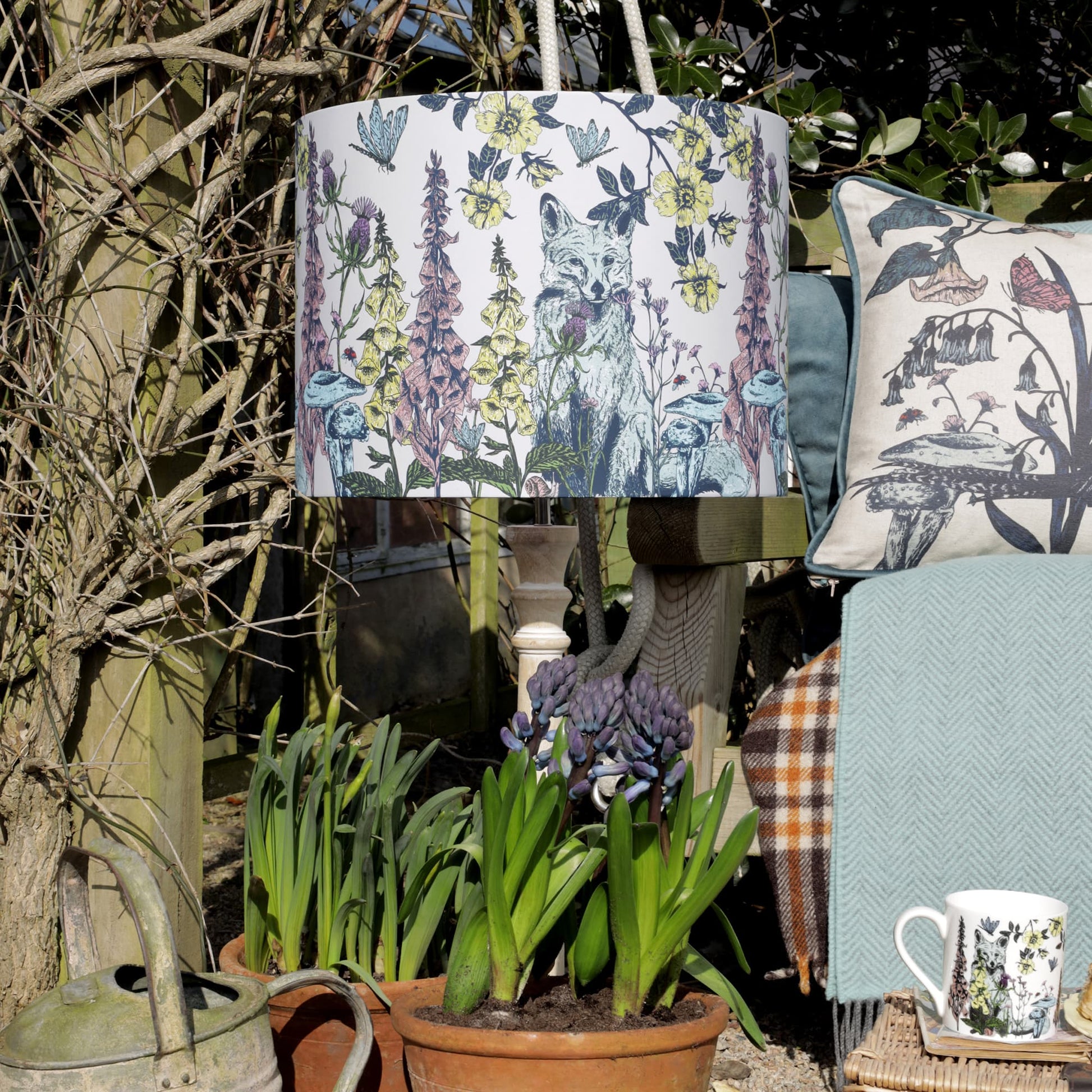 A nature-inspired outdoor setting featuring a botanical lampshade adorned with an intricate illustration of a fox among wildflowers, set on a rustic wooden lampstand. Surrounding the lamp are potted hyacinths, a vintage watering can, and a cozy seating area with patterned cushions and a herringbone throw, creating a charming and inviting garden retreat