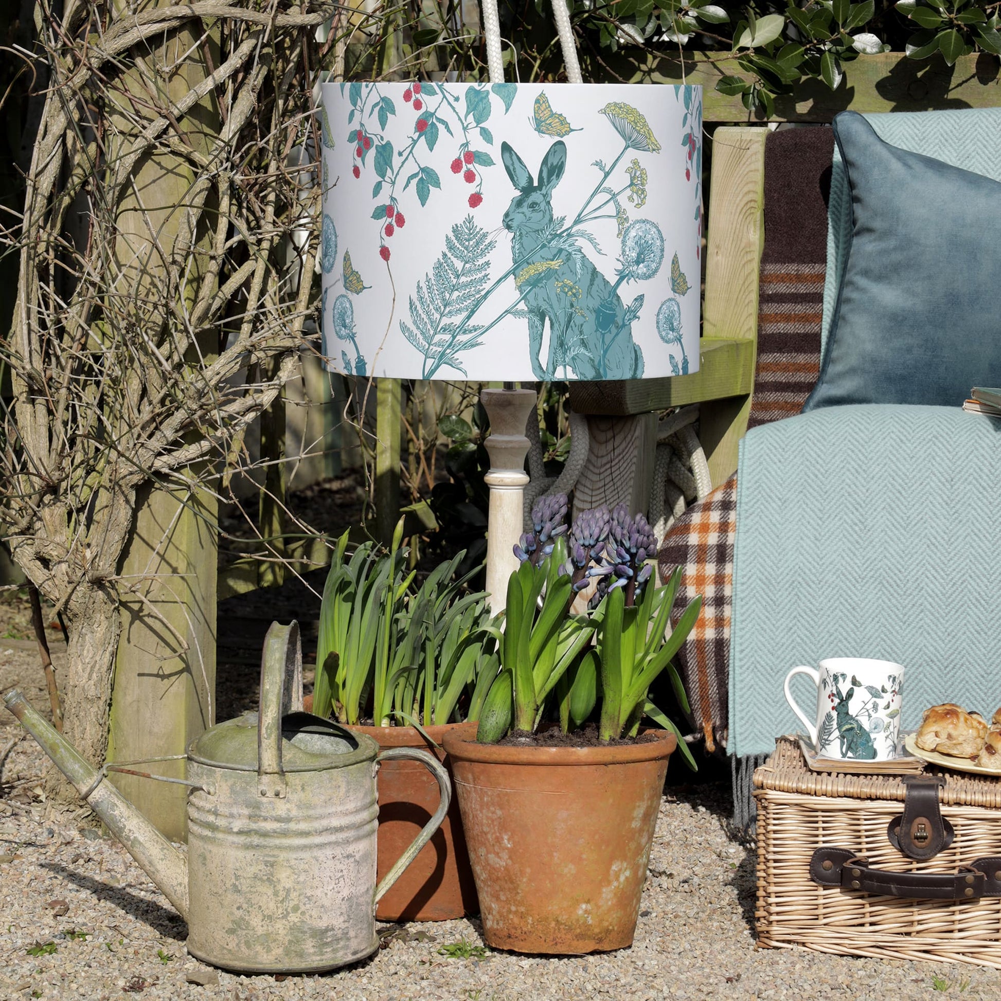 Charming outdoor setting with a botanical rabbit-print lampshade, a rustic watering can, and potted hyacinths. A cozy bench with a soft blue throw and velvet cushion sits nearby, with a wicker picnic basket holding a floral mug and scones, creating a peaceful countryside retreat.