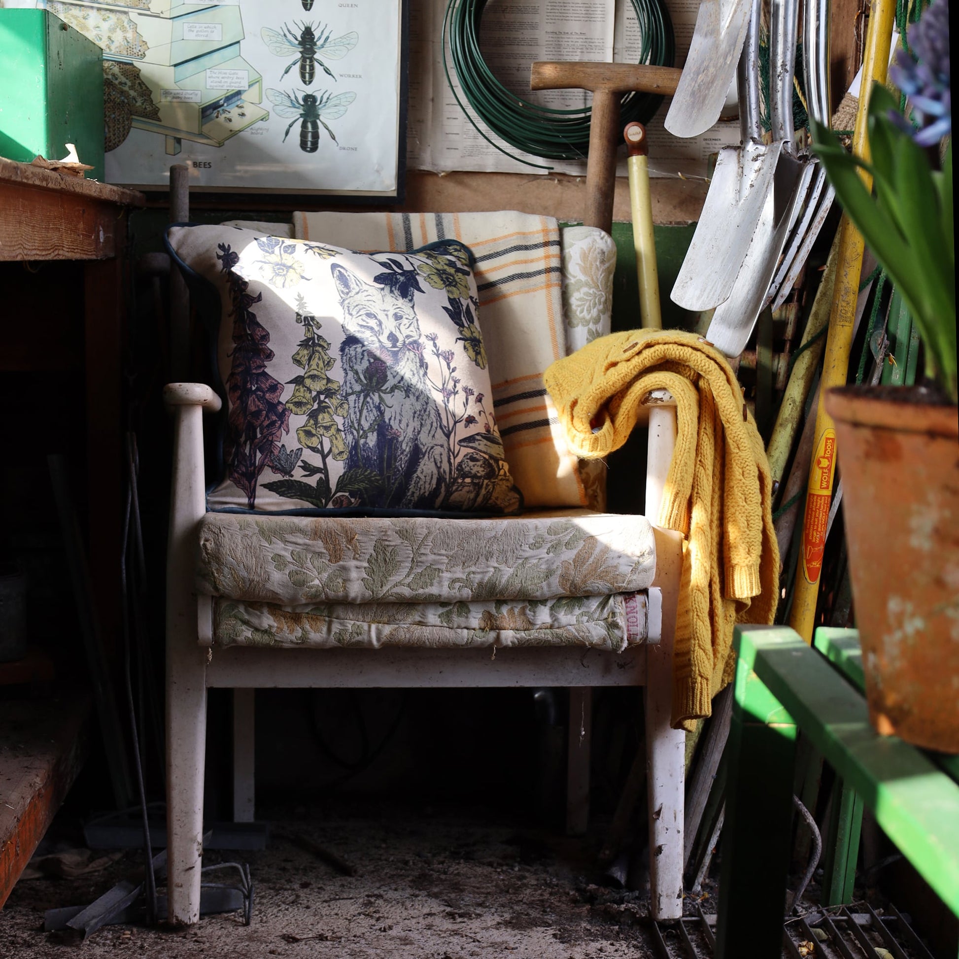 A cozy corner in a rustic garden shed featuring a vintage white wooden chair with a botanical and wildlife-themed cushion depicting a fox among wildflowers. A mustard yellow knitted sweater is draped over the chair’s armrest. The space is surrounded by gardening tools, potted plants, and warm sunlight filtering in, creating an inviting and nostalgic atmosphere.